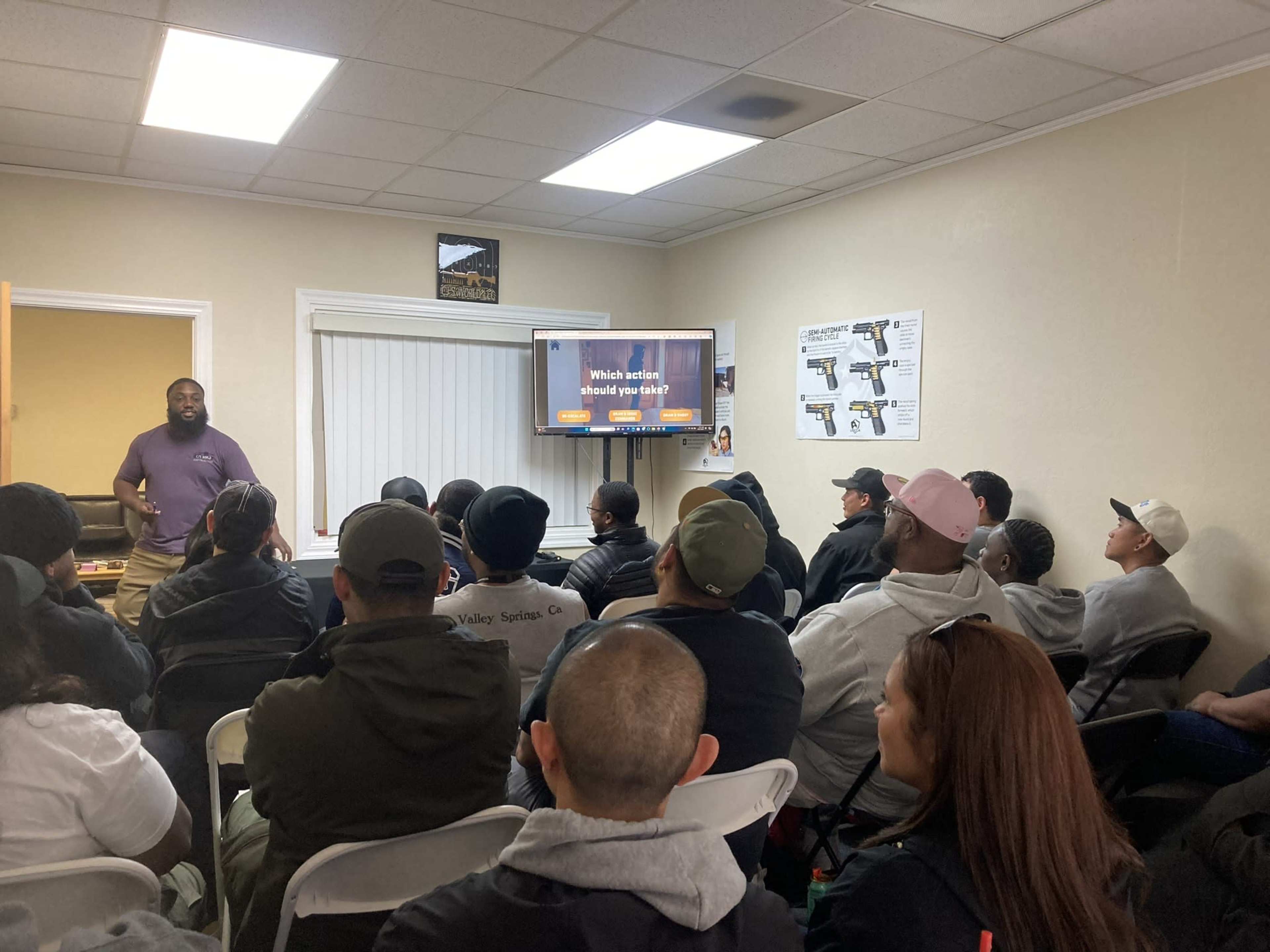 A diverse group of individuals sits in a room facing a speaker and a television displaying a presentation.