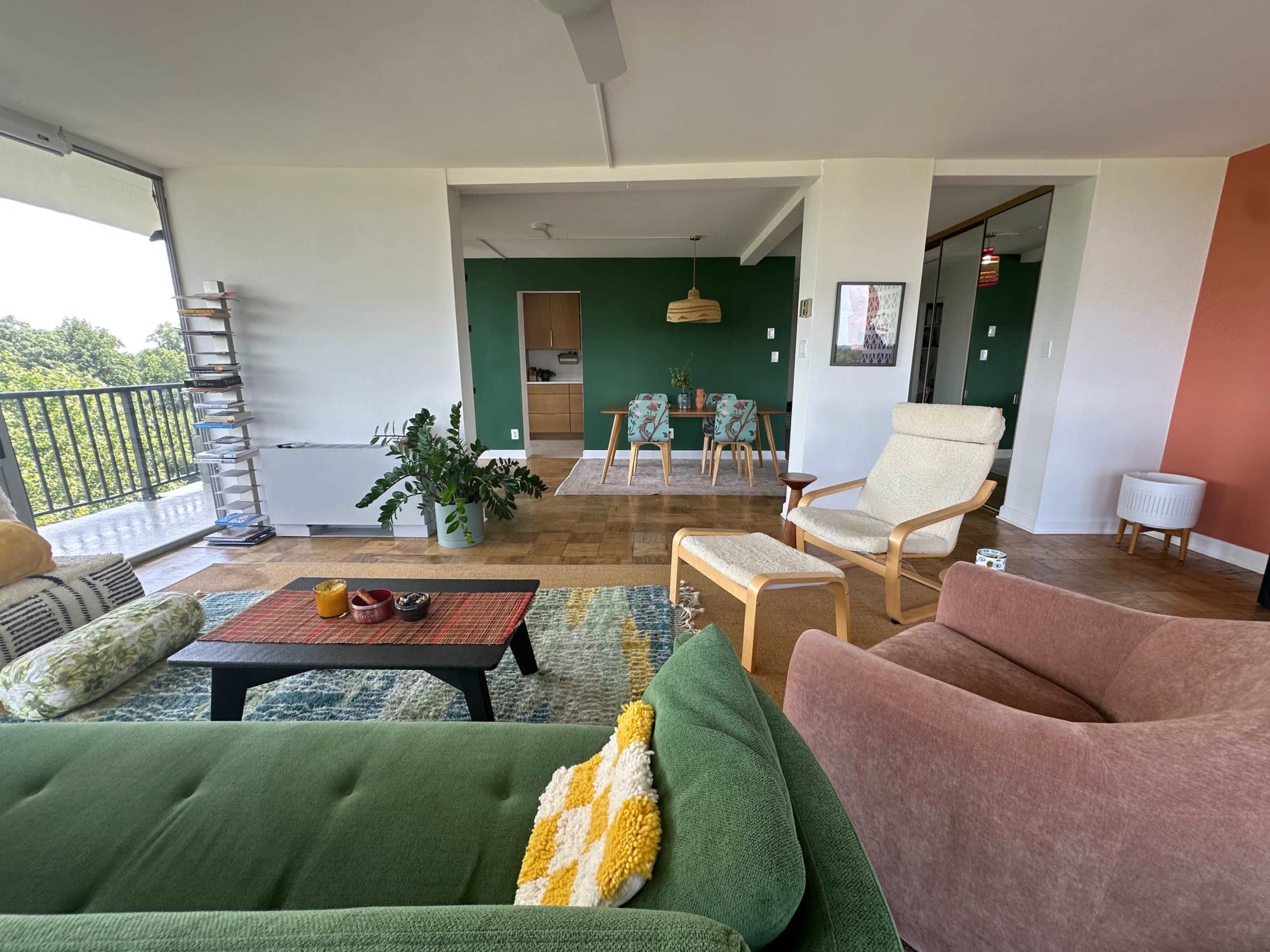 The image shows a bright, modern living room with a green sofa, a brown armchair, a coffee table with decorative items, and a dining area in the background.