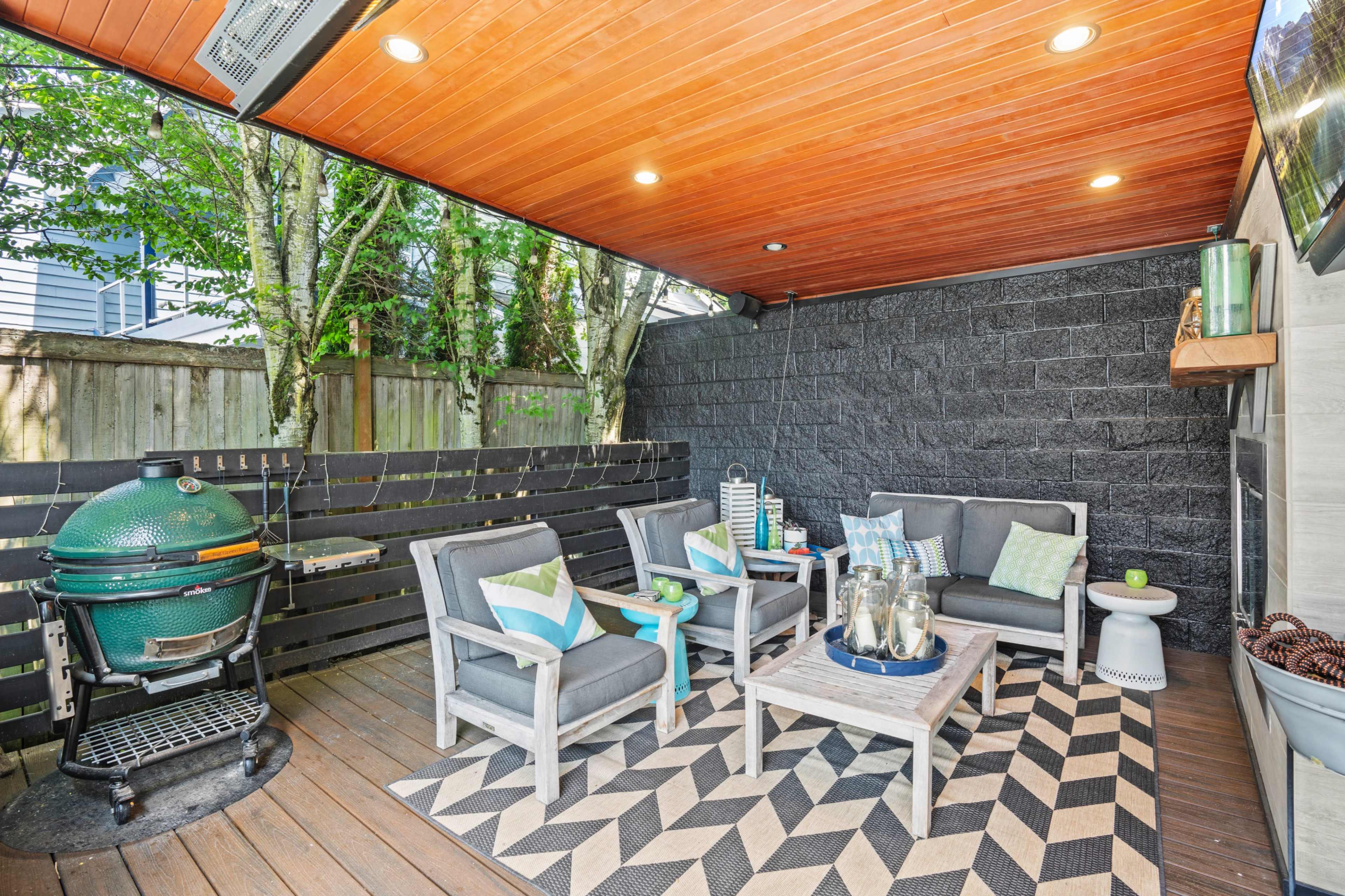 A covered outdoor seating area with gray chairs, a coffee table, and a barbecue grill against a backdrop of wooden fencing and trees.