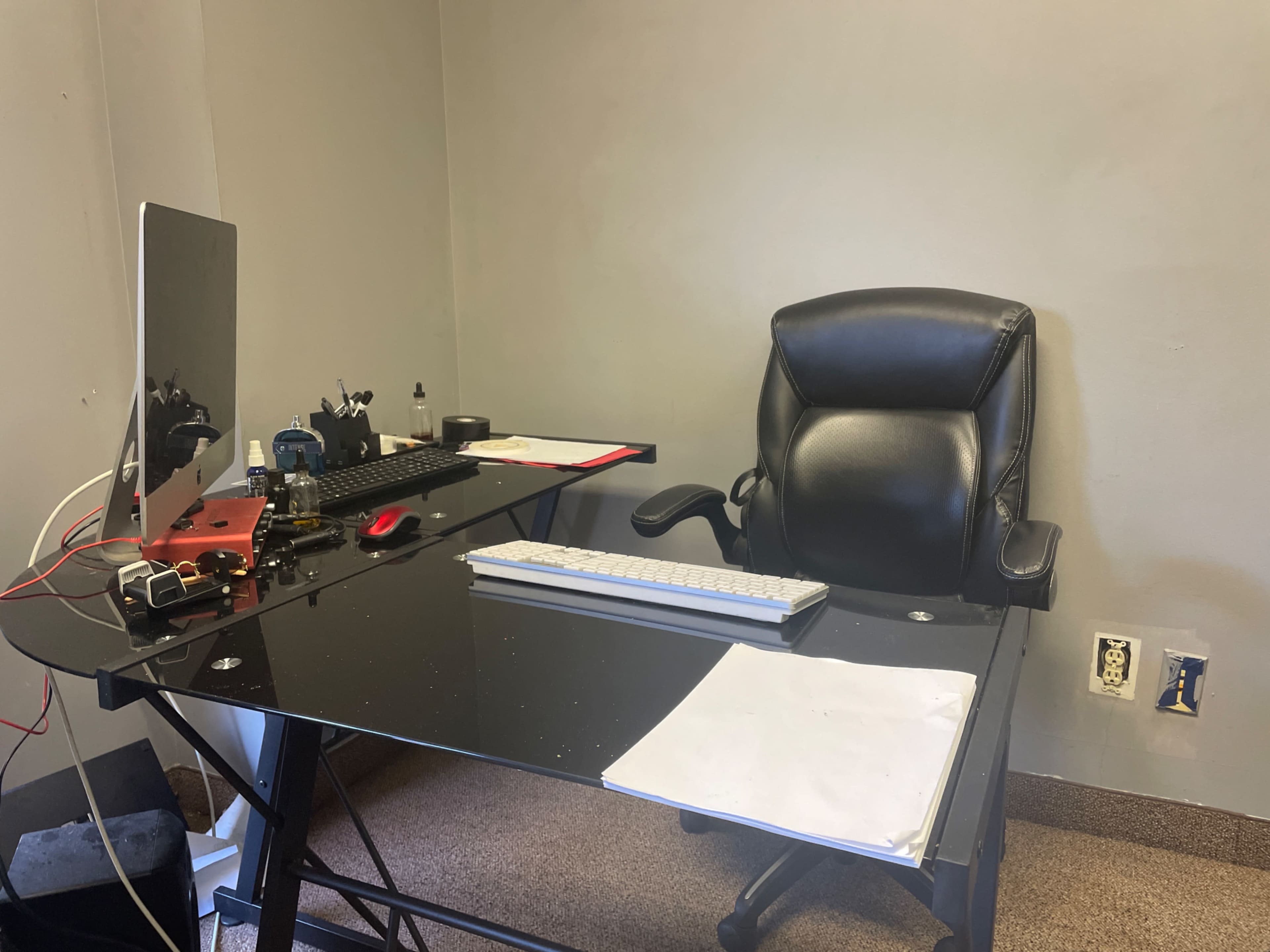 A black office chair next to a glass desk equipped with a computer monitor, keyboard, and some scattered papers.