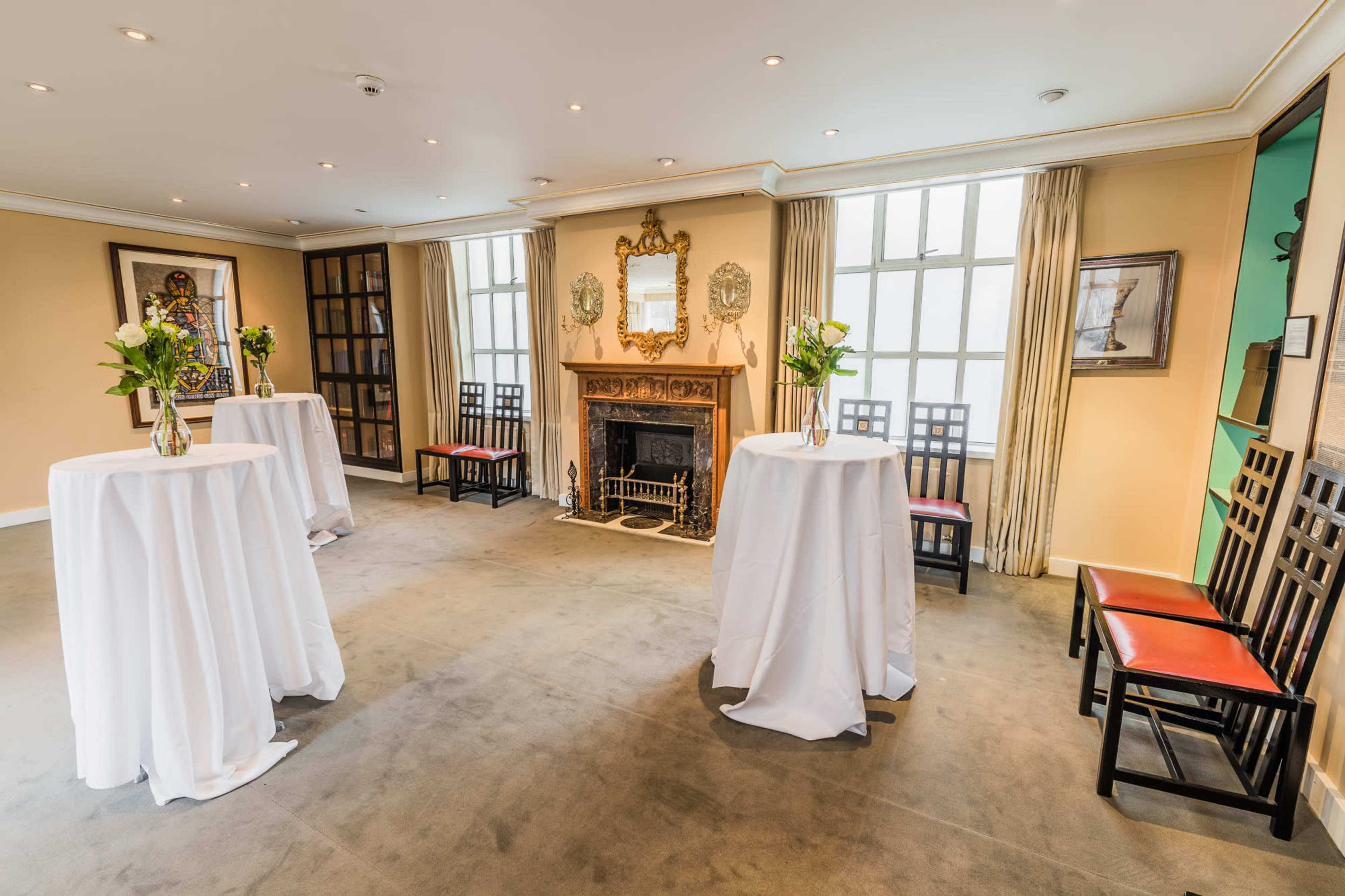 A well-lit event room with four white-draped tables, a fireplace, and wooden chairs arranged around the space.