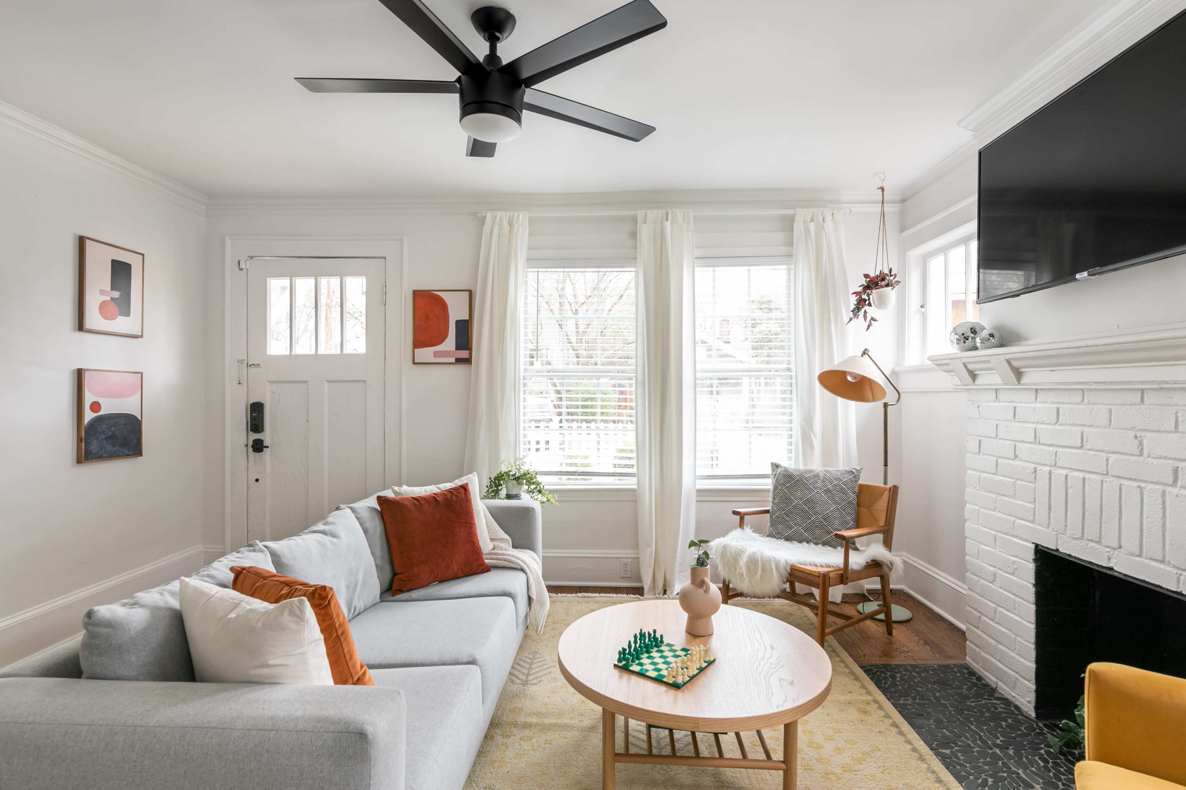 A modern living room with a light gray sofa, a round wooden coffee table, and a fireplace, featuring a door leading outside and large windows with white curtains.