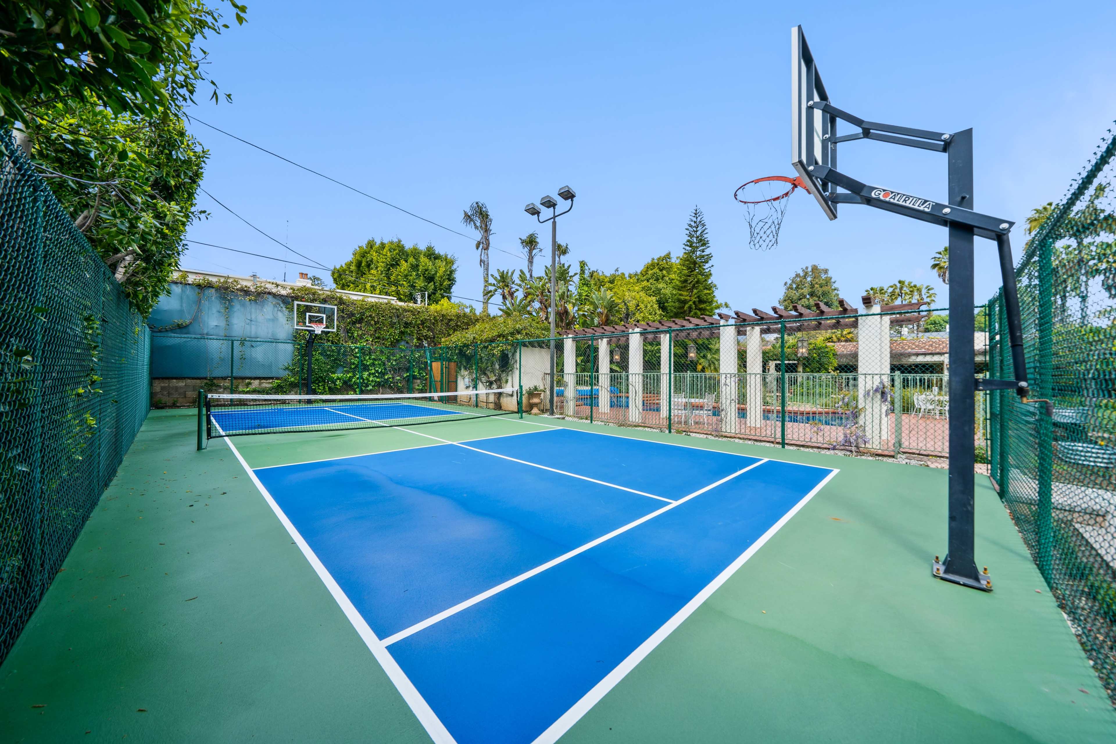 The image shows a blue and green basketball court surrounded by a fence and greenery, with a hoop at one end.