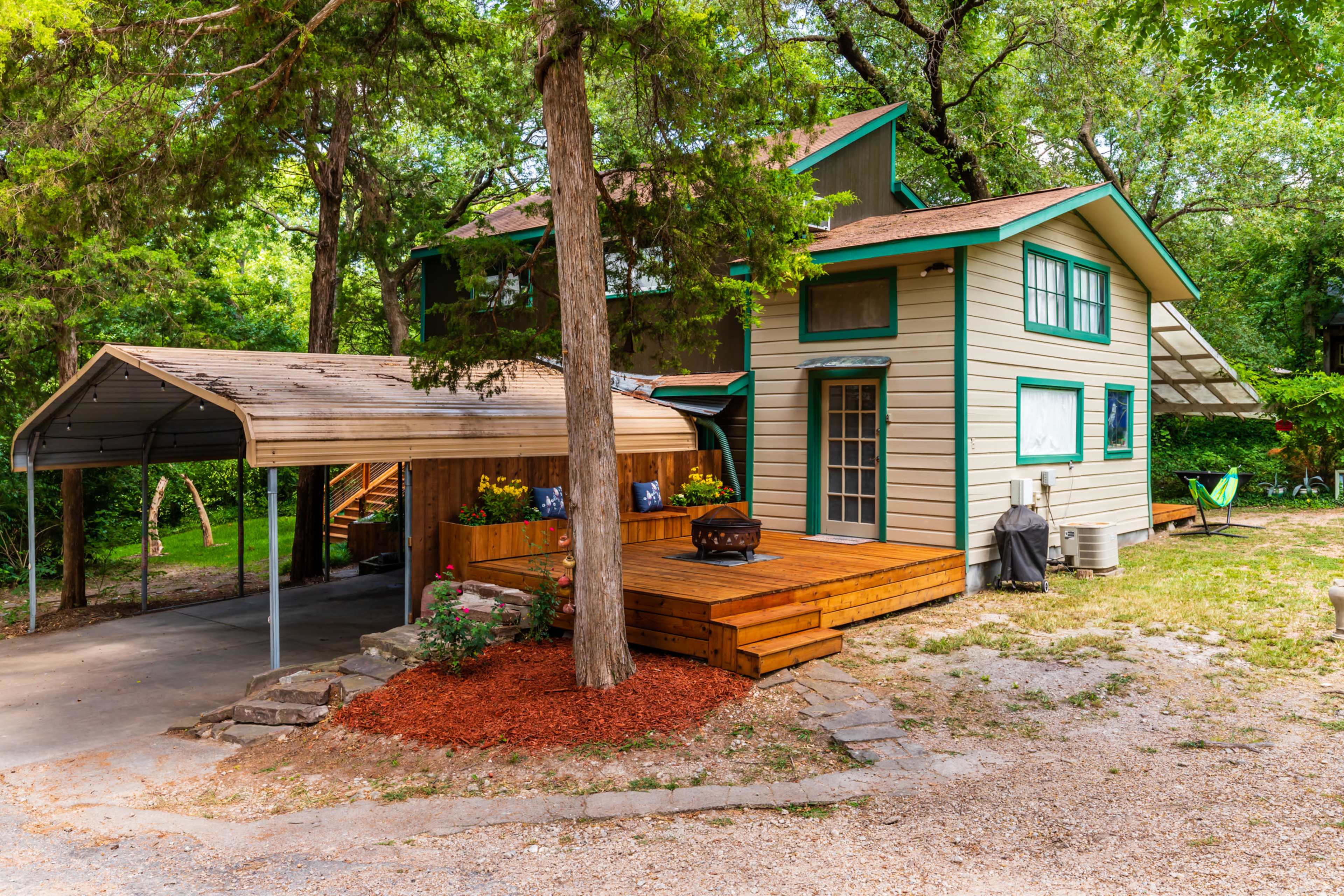A two-story house with a green and beige exterior features a large wooden deck, a carport, and a landscaped yard surrounded by trees.
