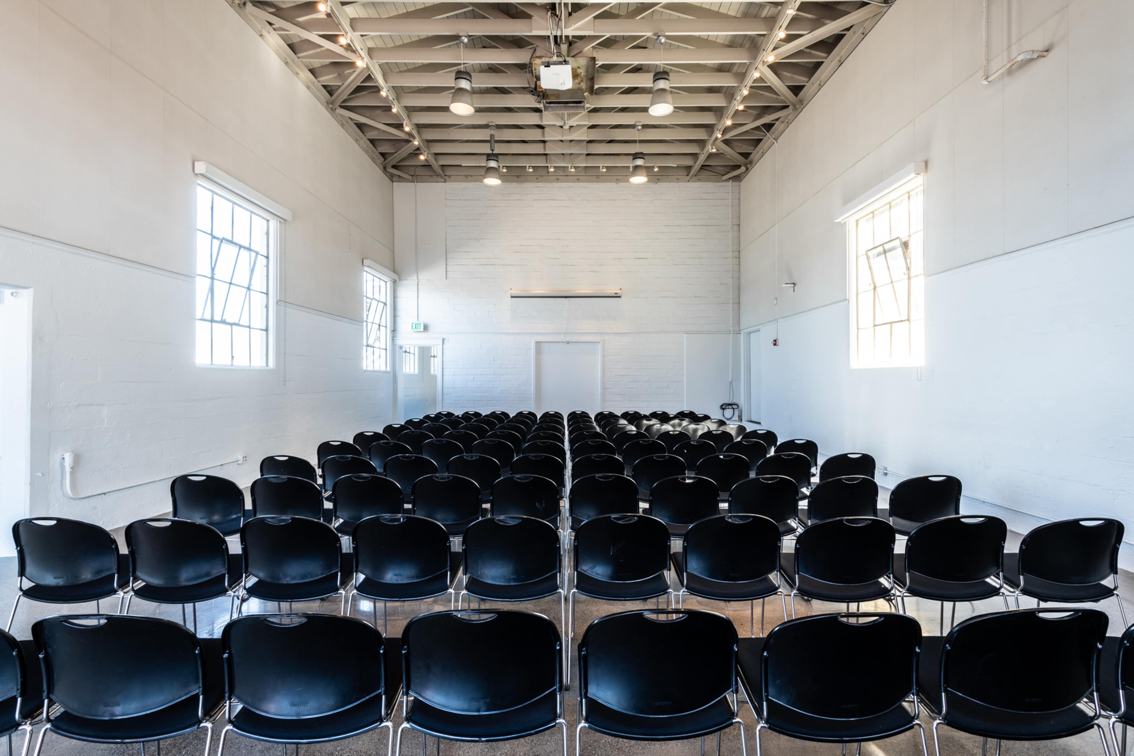 A row of black chairs is arranged in neat rows in a bright, empty hall with large windows.