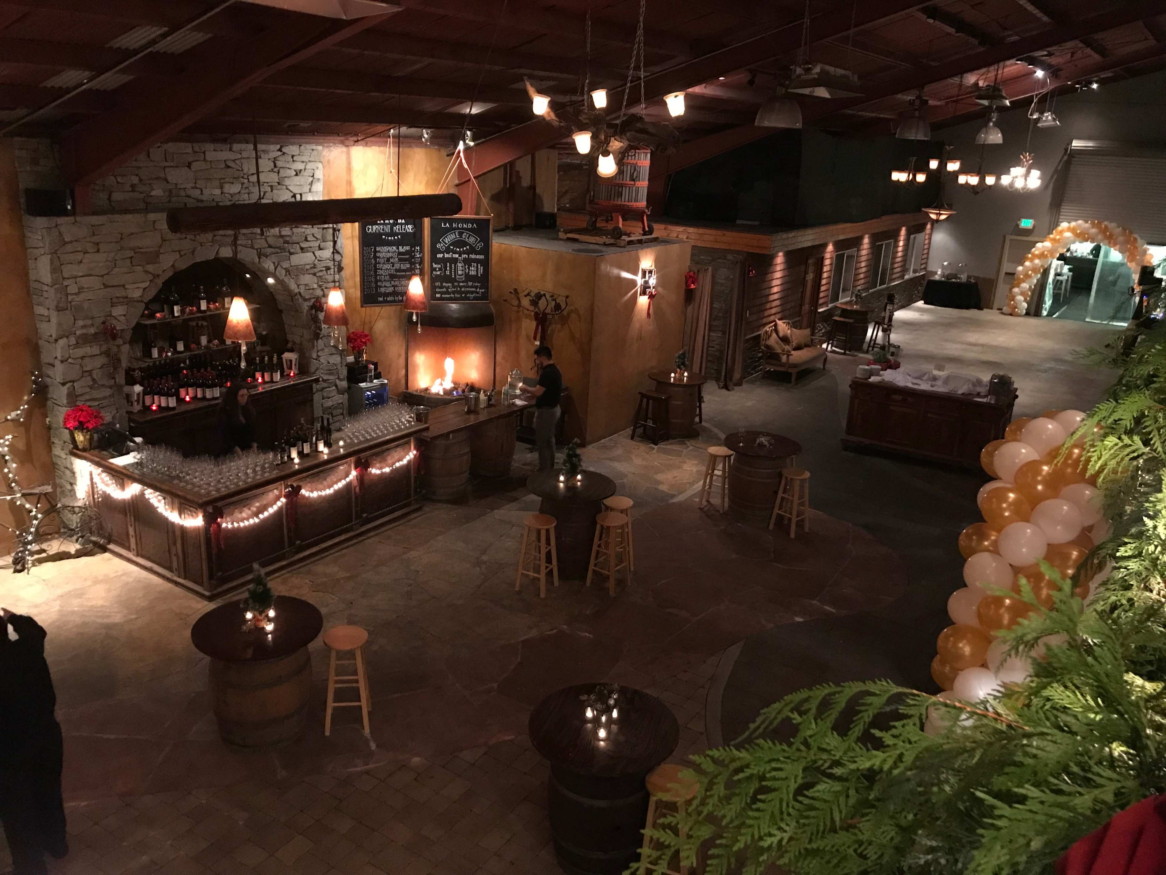 An indoor event space featuring a bar made of wood and stone, with tables arranged around the area and decorative lighting overhead.