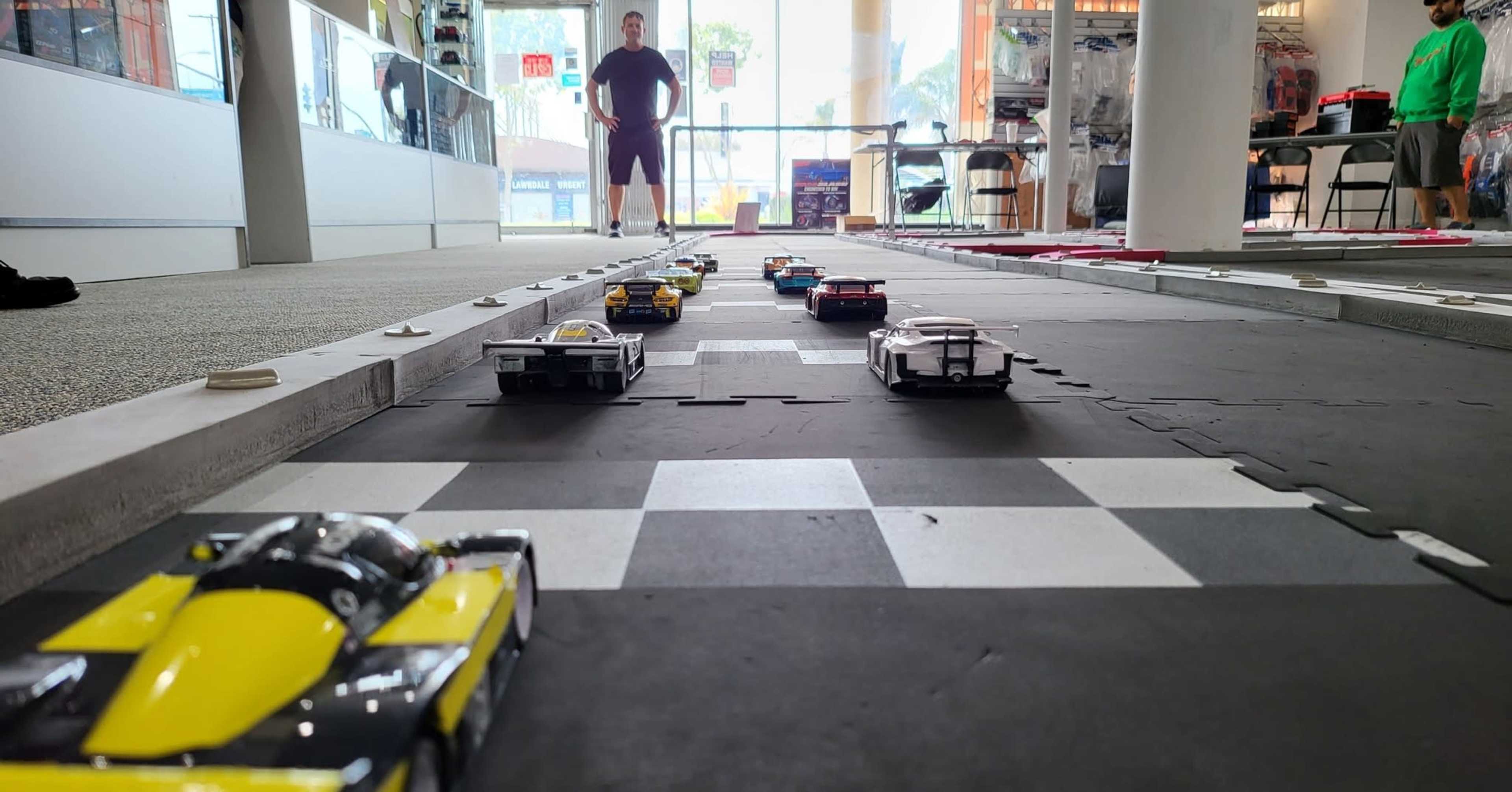 A line of toy race cars moves along a black and white checkered track in a store while a person watches from the background.