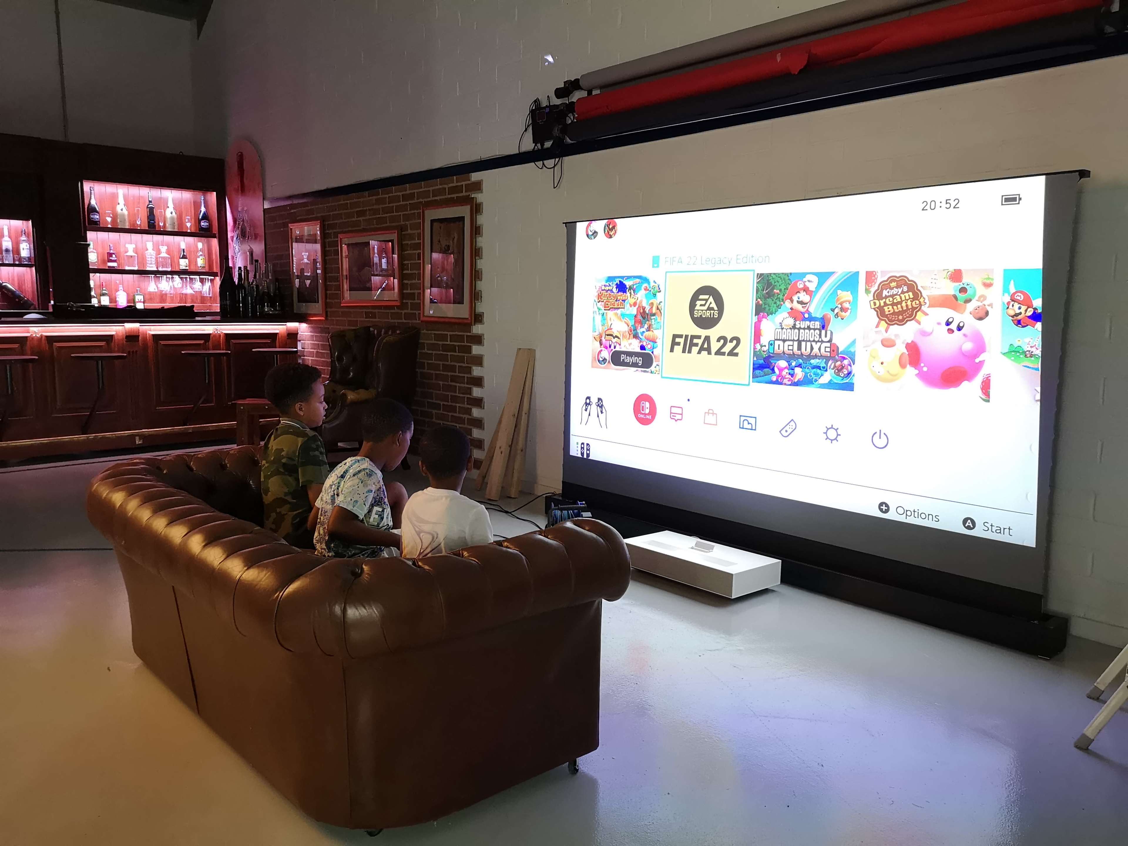 Two children sit on a brown couch playing video games on a large projector screen in a casual indoor setting.