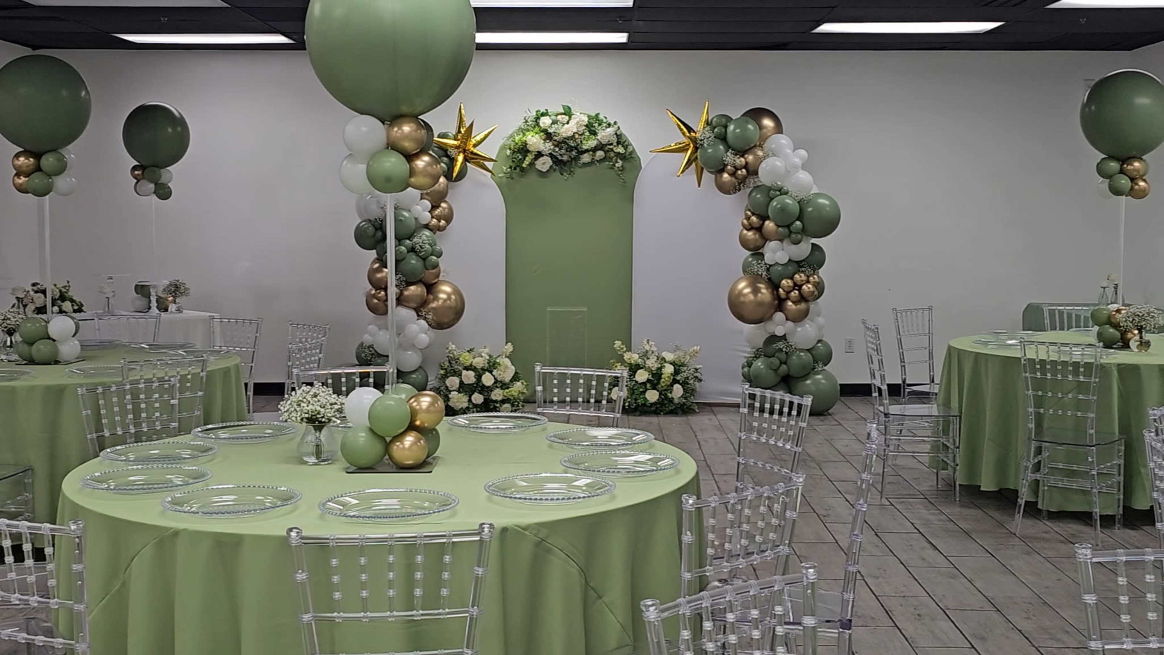 The image shows a banquet hall decorated with green and gold balloons, floral arrangements, and set with round tables featuring clear chairs.