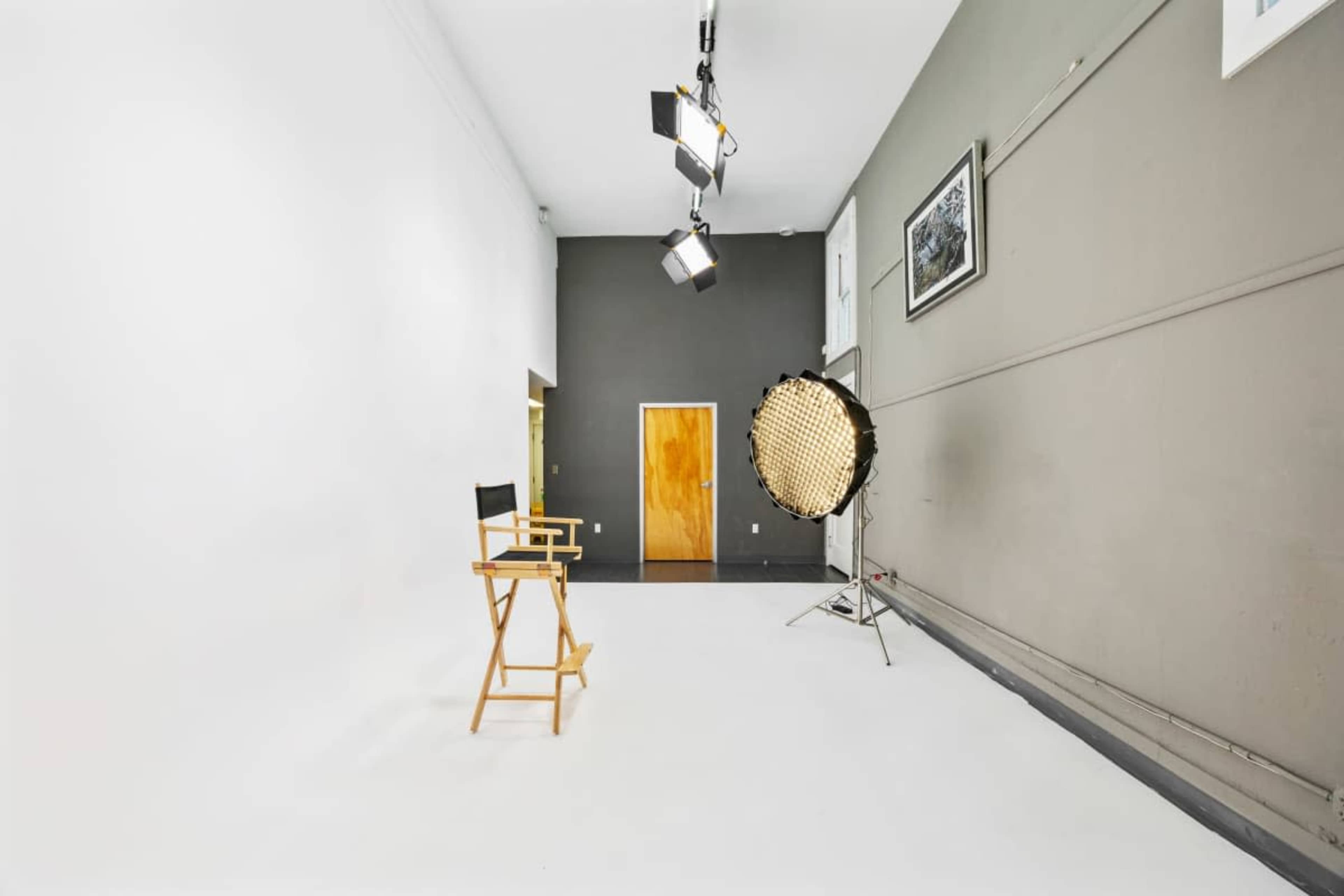 The image shows a photography studio featuring a wooden director's chair, a large light source on a stand, and a plain white wall.