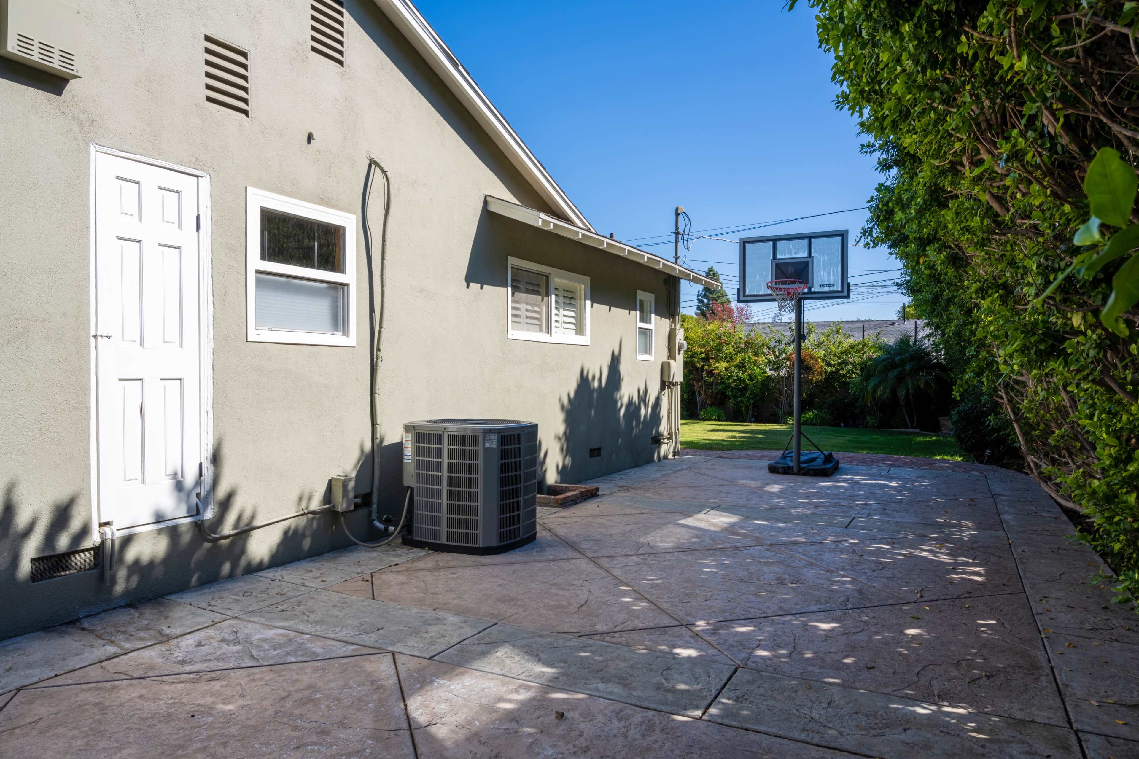 A backyard patio with a basketball hoop, an air conditioning unit, and a house wall surrounded by greenery.