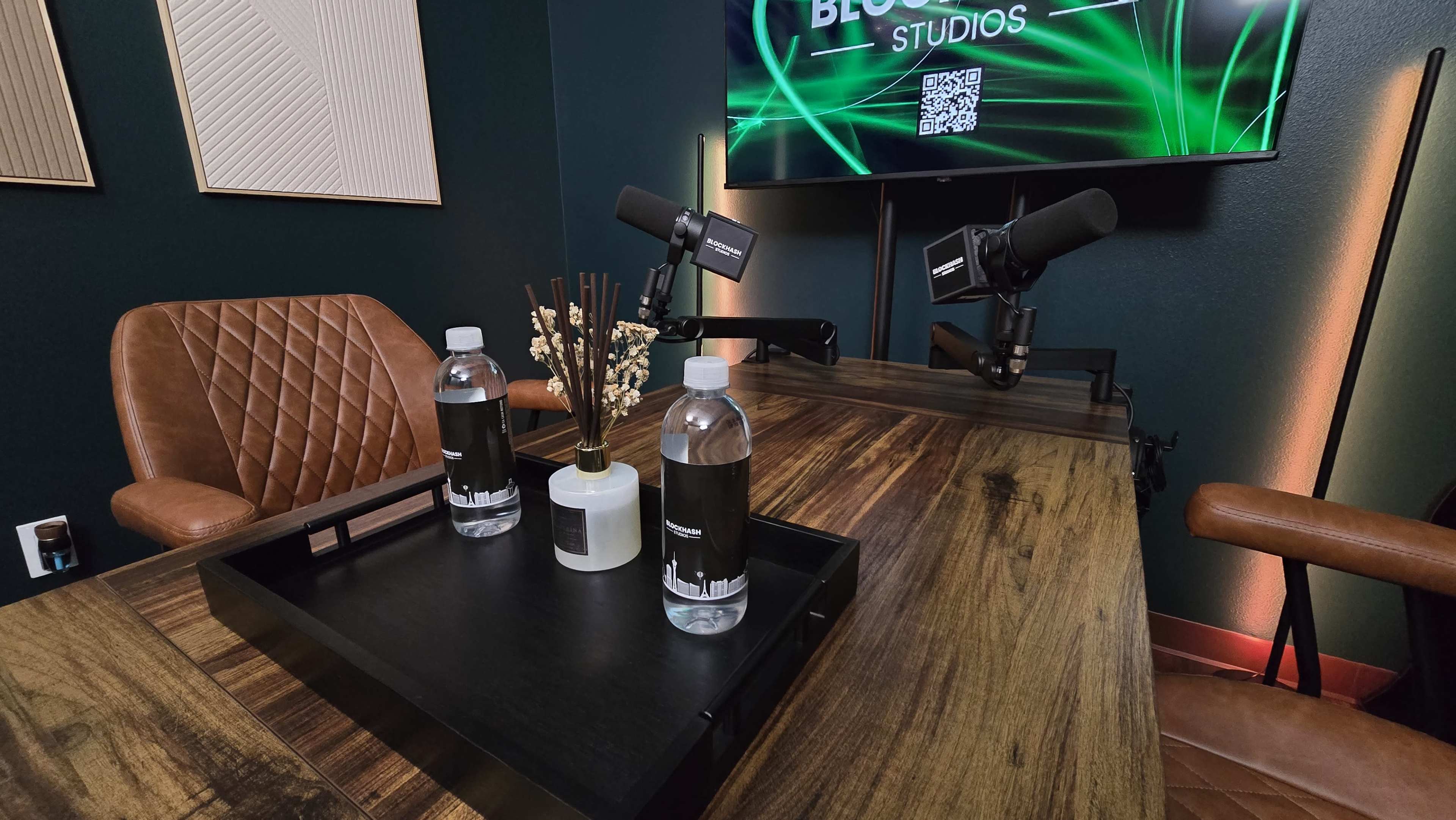 The image shows a modern studio setup with a wooden table featuring two water bottles, a decorative vase, and two microphones, against a backdrop of green lighting and a screen displaying "BLOOM STUDIOS."