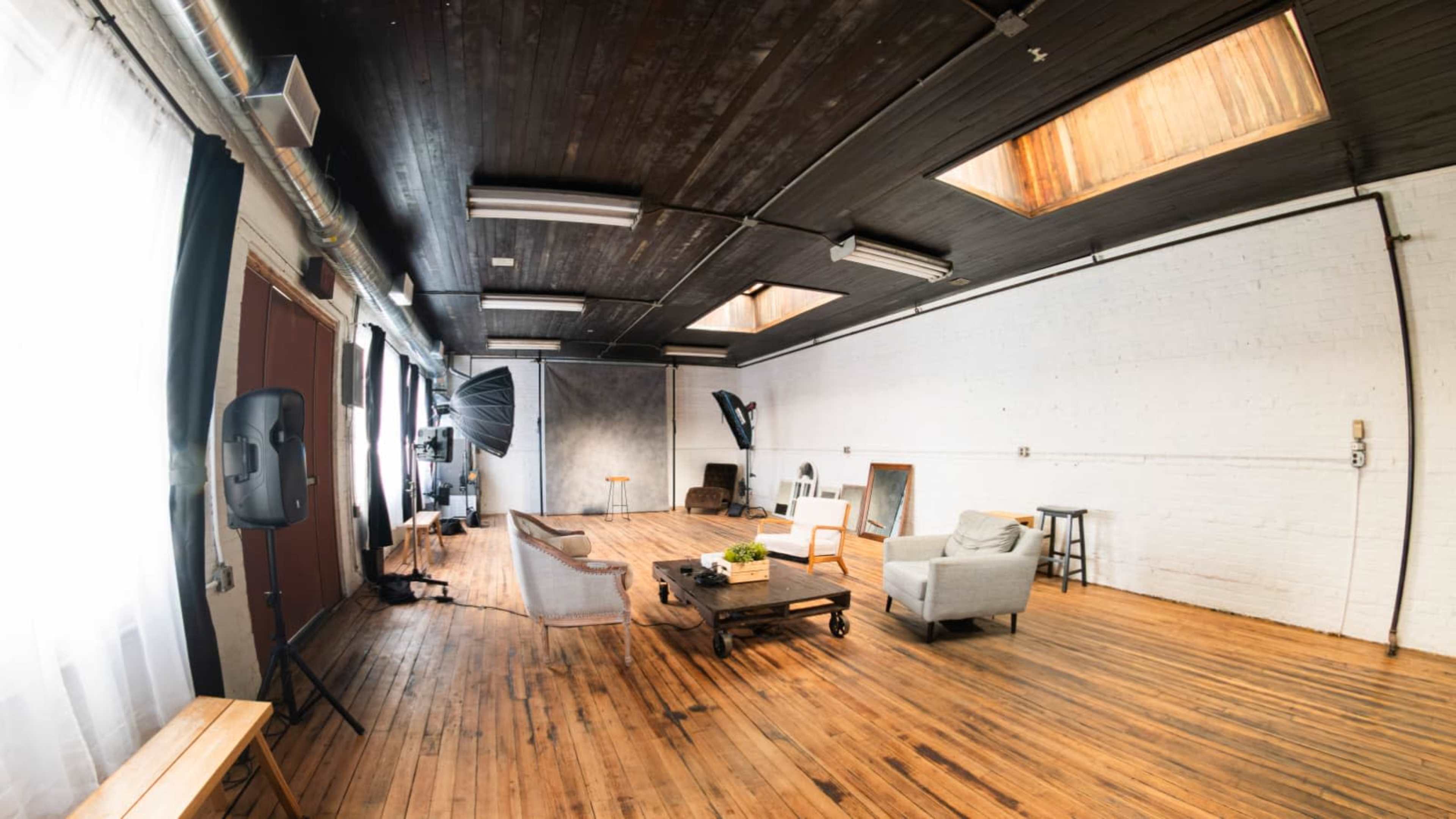The image shows a spacious photography studio featuring wooden floors, large windows, and minimalist furniture arranged in a well-lit environment.