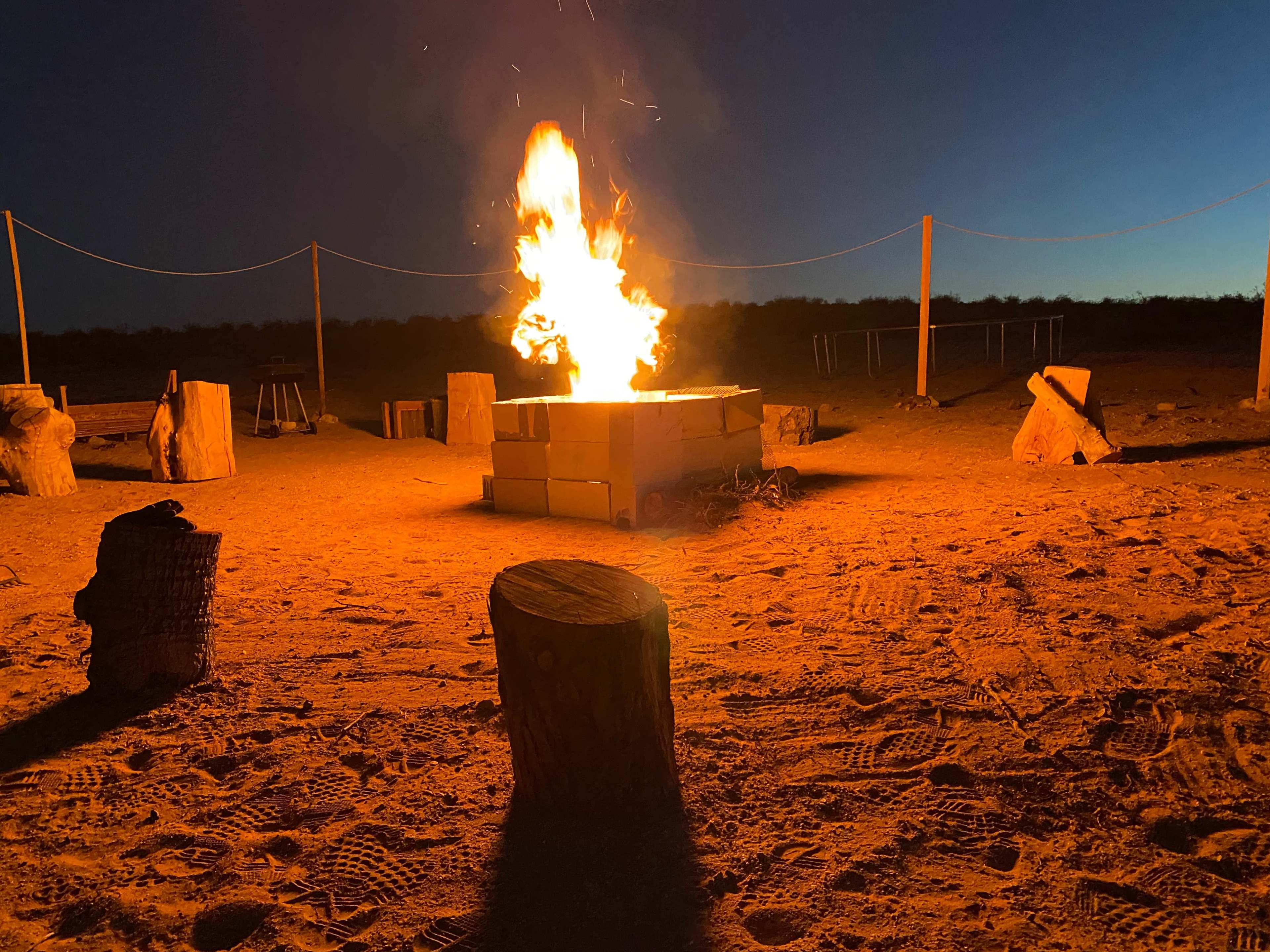 A campfire blazes at dusk, surrounded by wooden logs and sand.