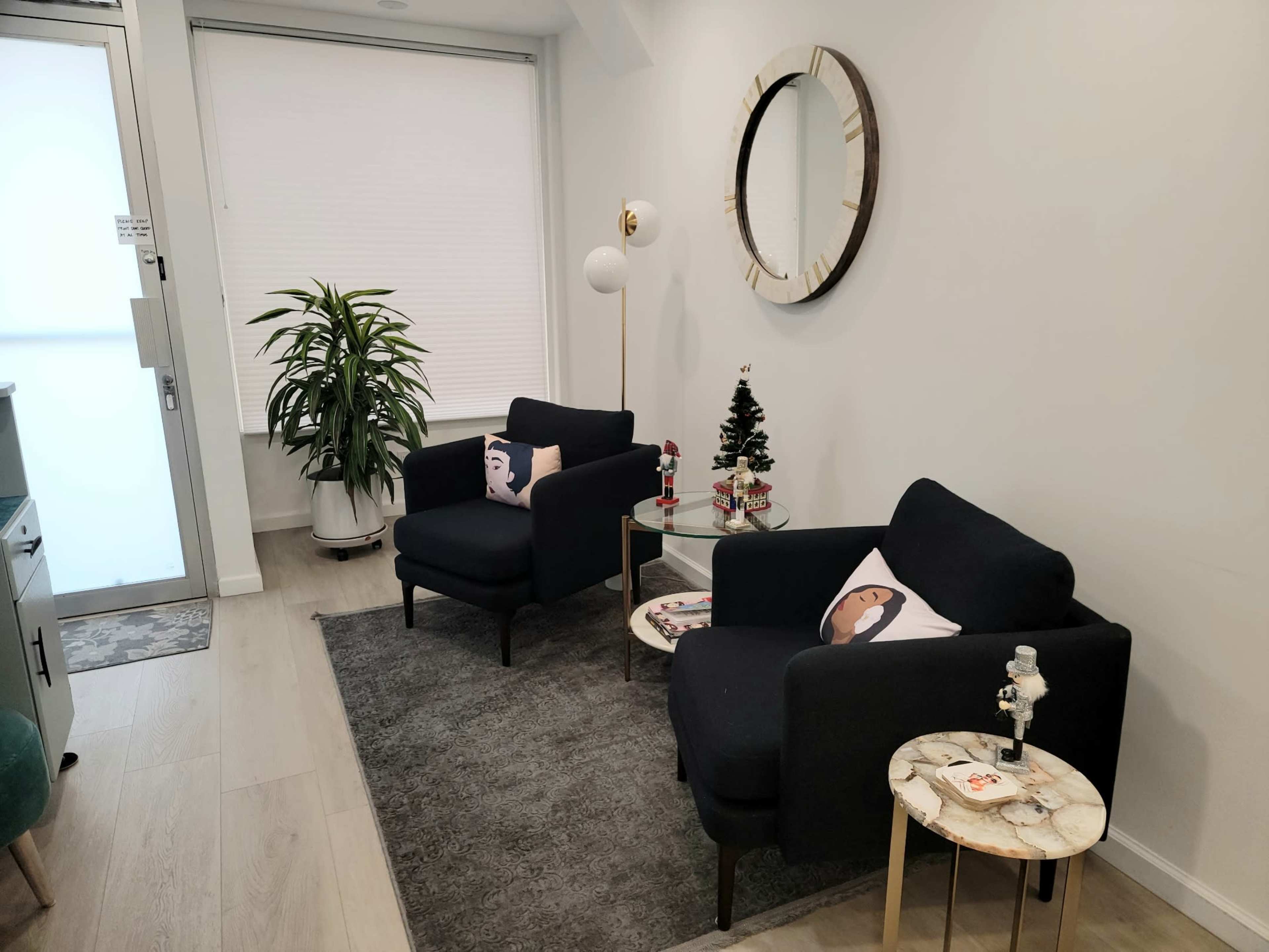 A welcoming interior space features two black armchairs with decorative pillows, a circular mirror, a small Christmas tree, and a potted plant, all arranged on a light wooden floor.