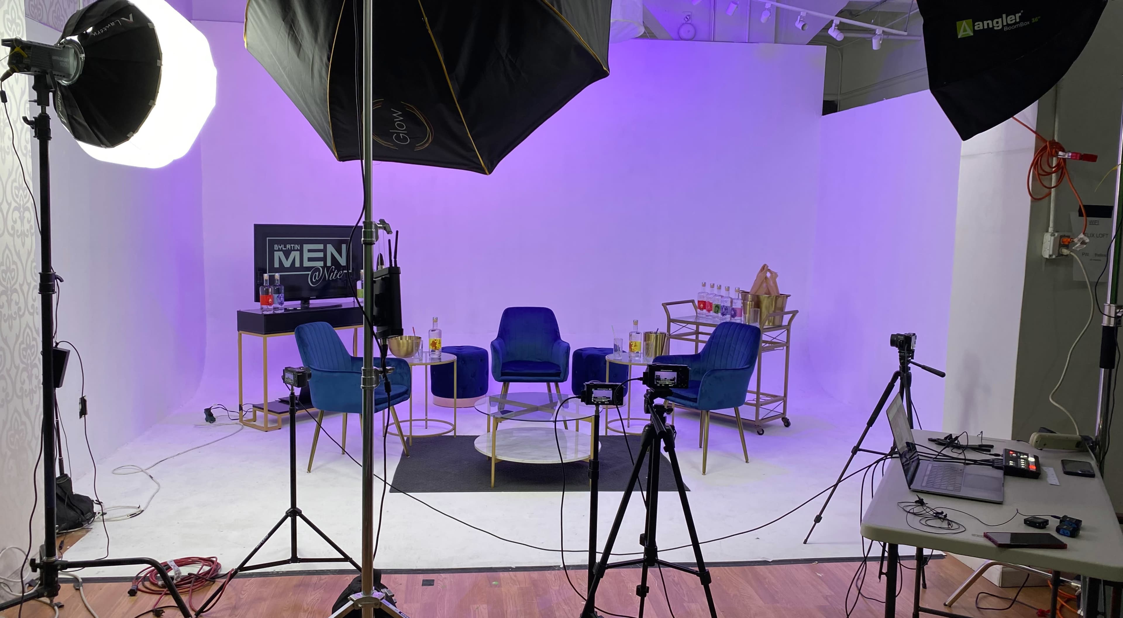 A studio setup with four blue chairs arranged around a glass coffee table, with lighting equipment and cameras positioned nearby.