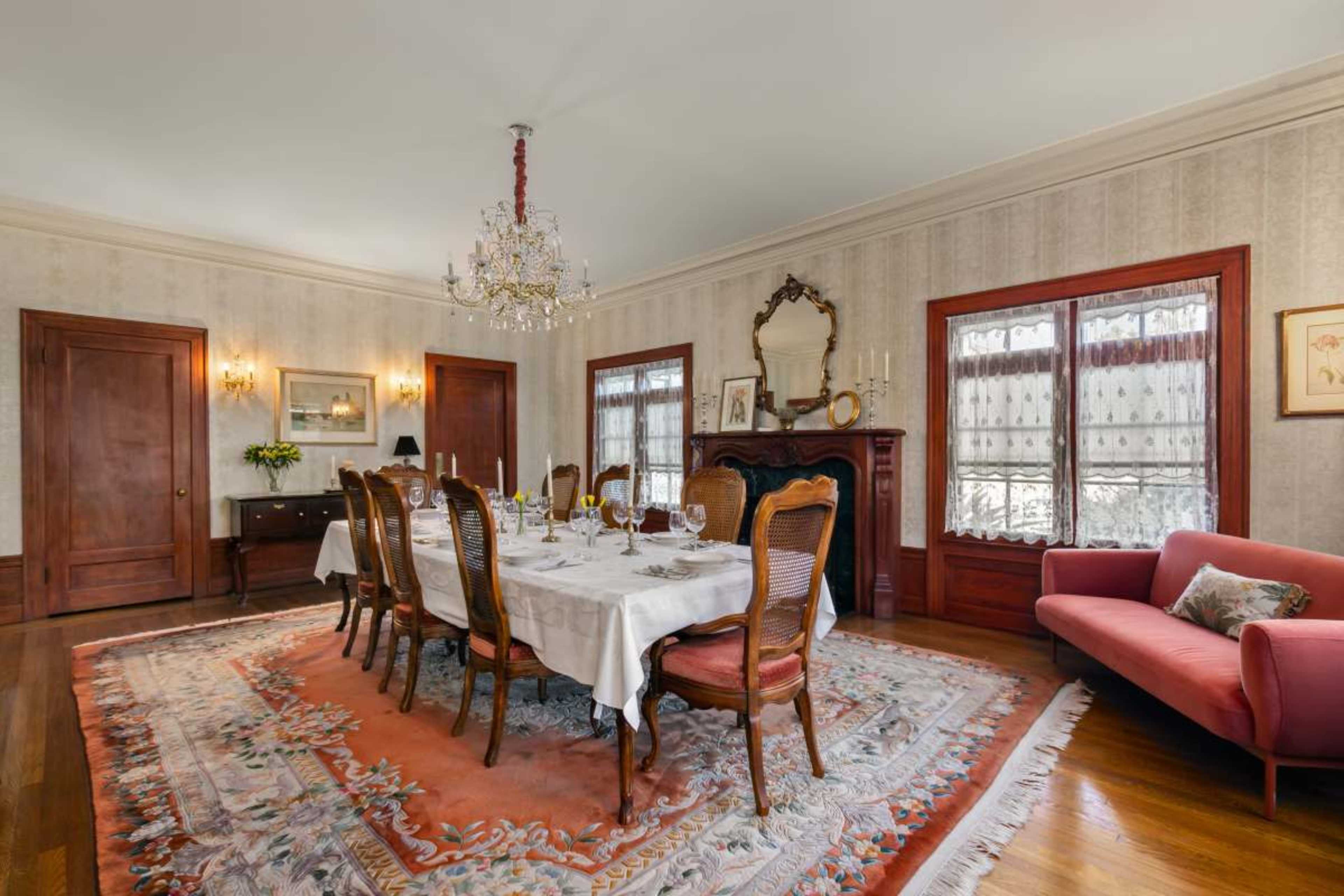 The image shows a dining room with a large table set for a meal, surrounded by chairs, a chandelier overhead, and a decorative rug on the wooden floor.
