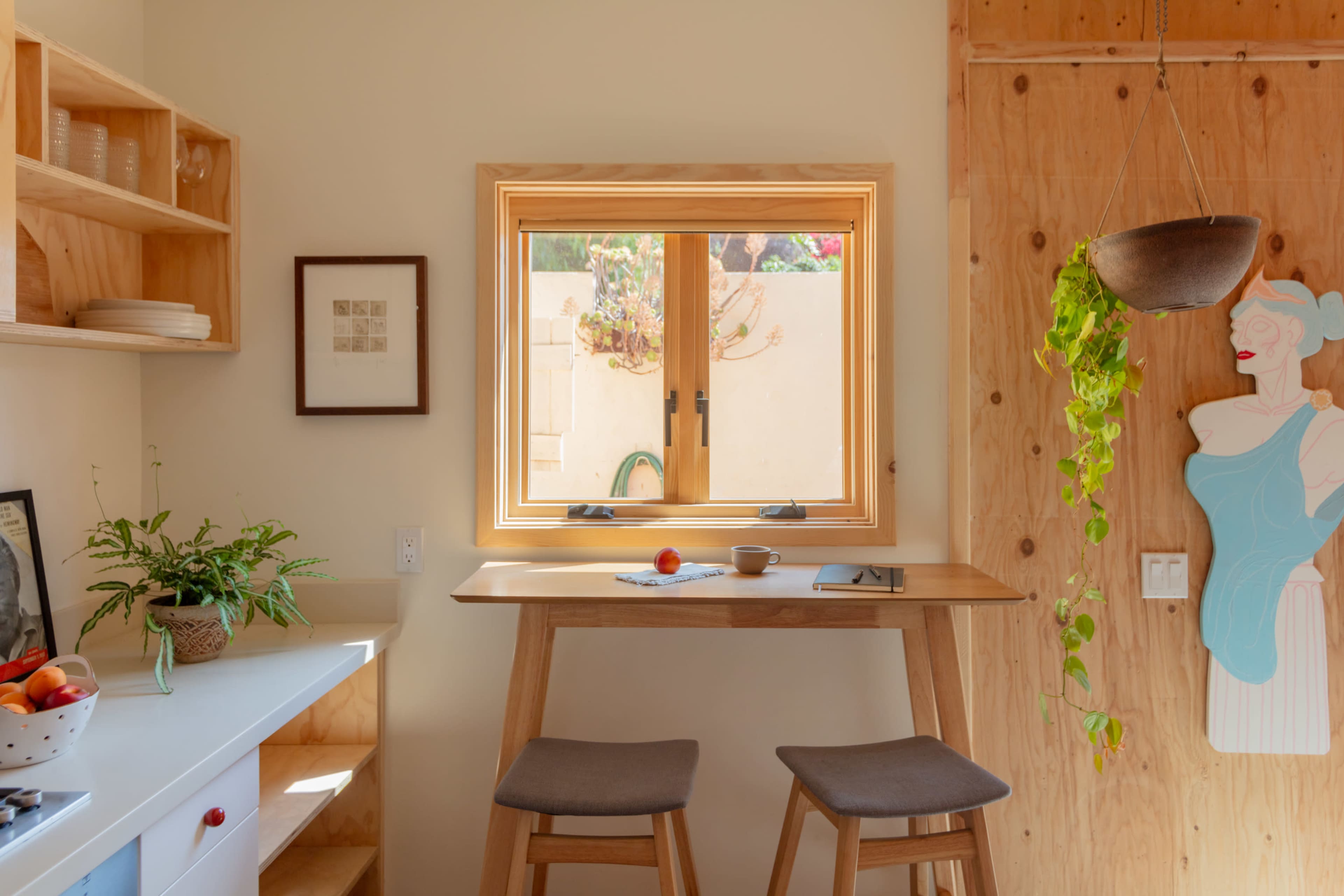 The image shows a cozy kitchen nook with a small table and two stools beneath a window, adorned with a potted plant and a few kitchen items.