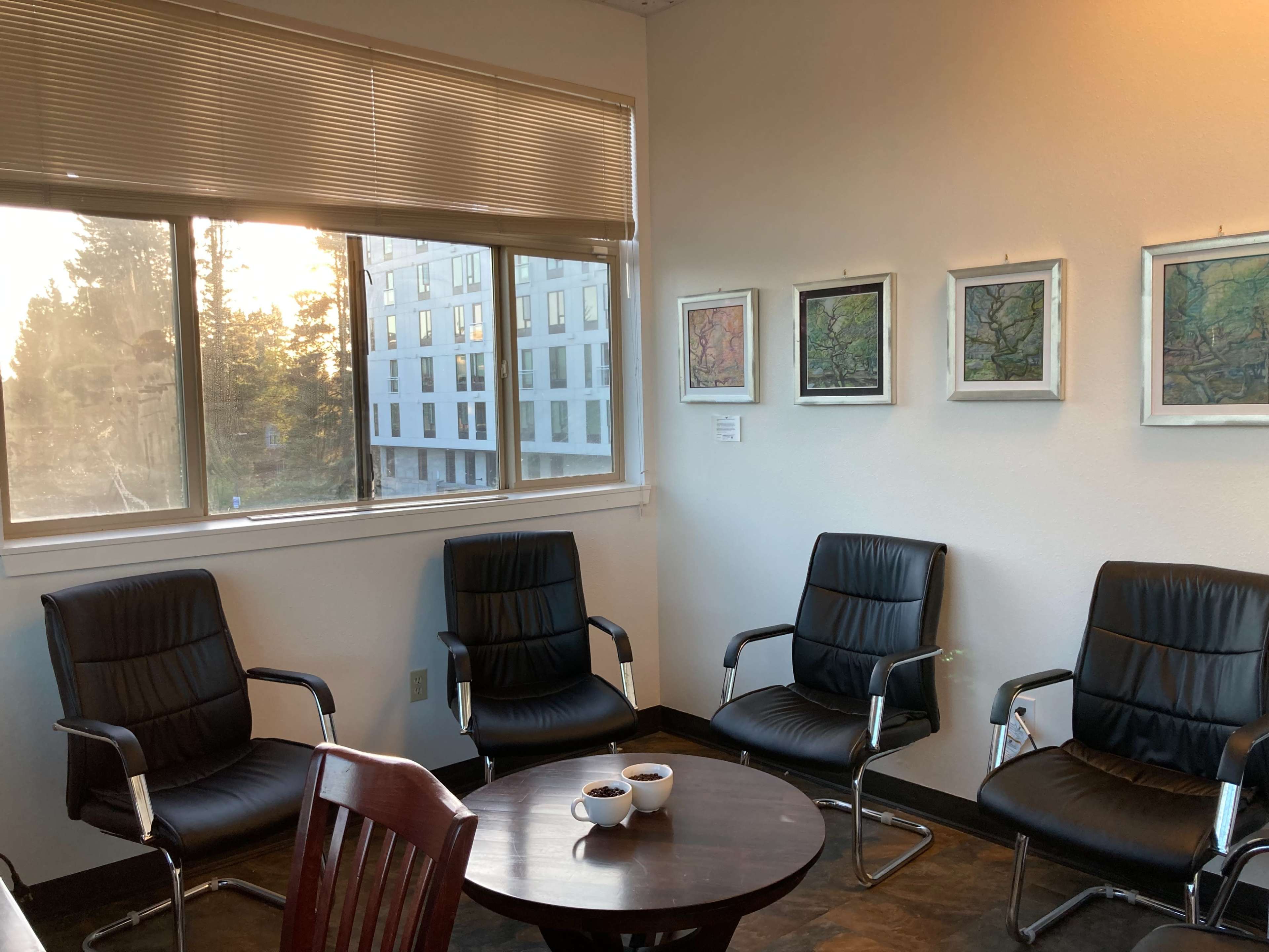 A waiting room features four black chairs surrounding a round table with two cups, and colorful artwork displayed on the walls near a window.