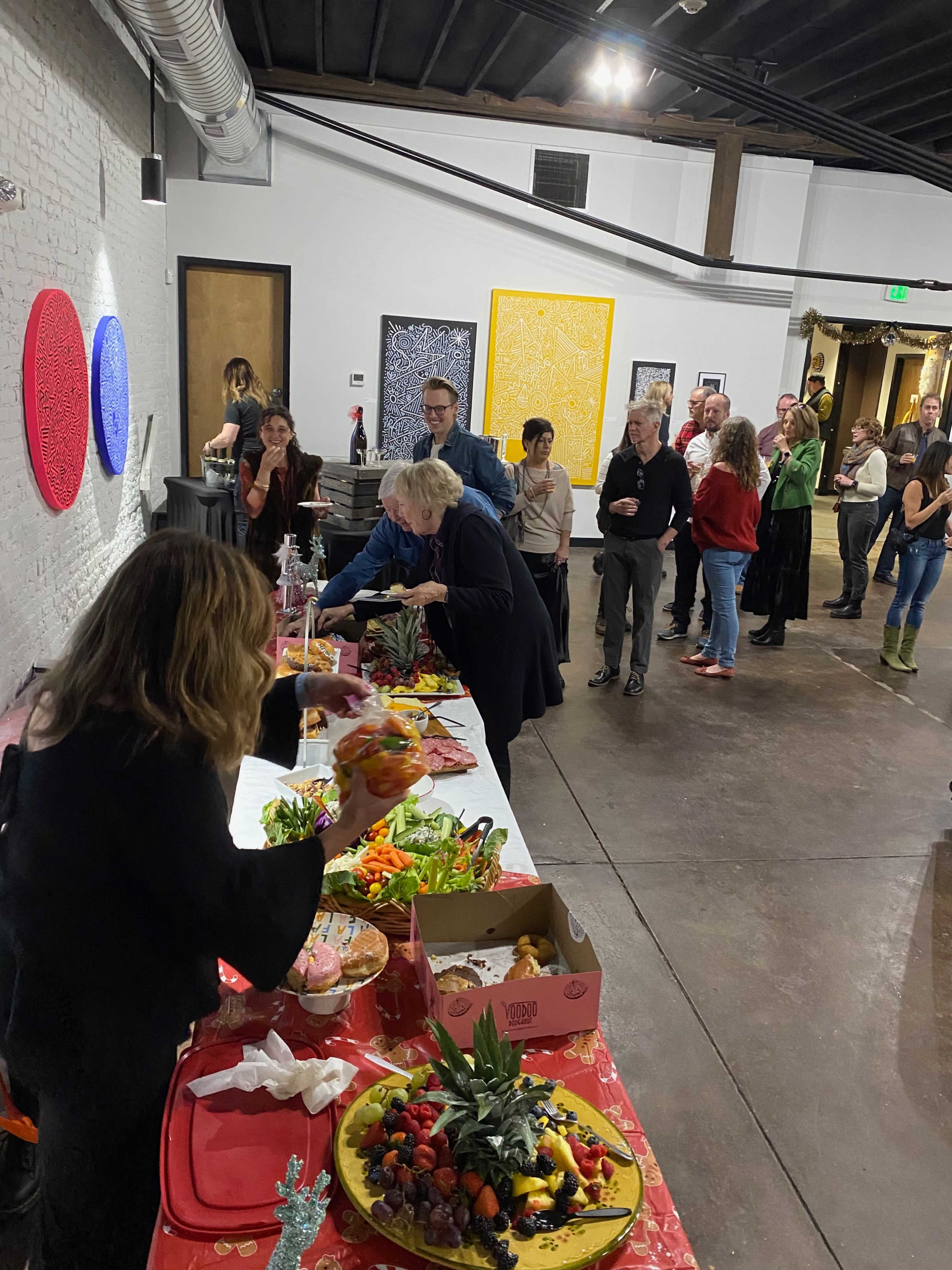 A group of people gathers in an art gallery while two individuals prepare a colorful array of food on a long table.