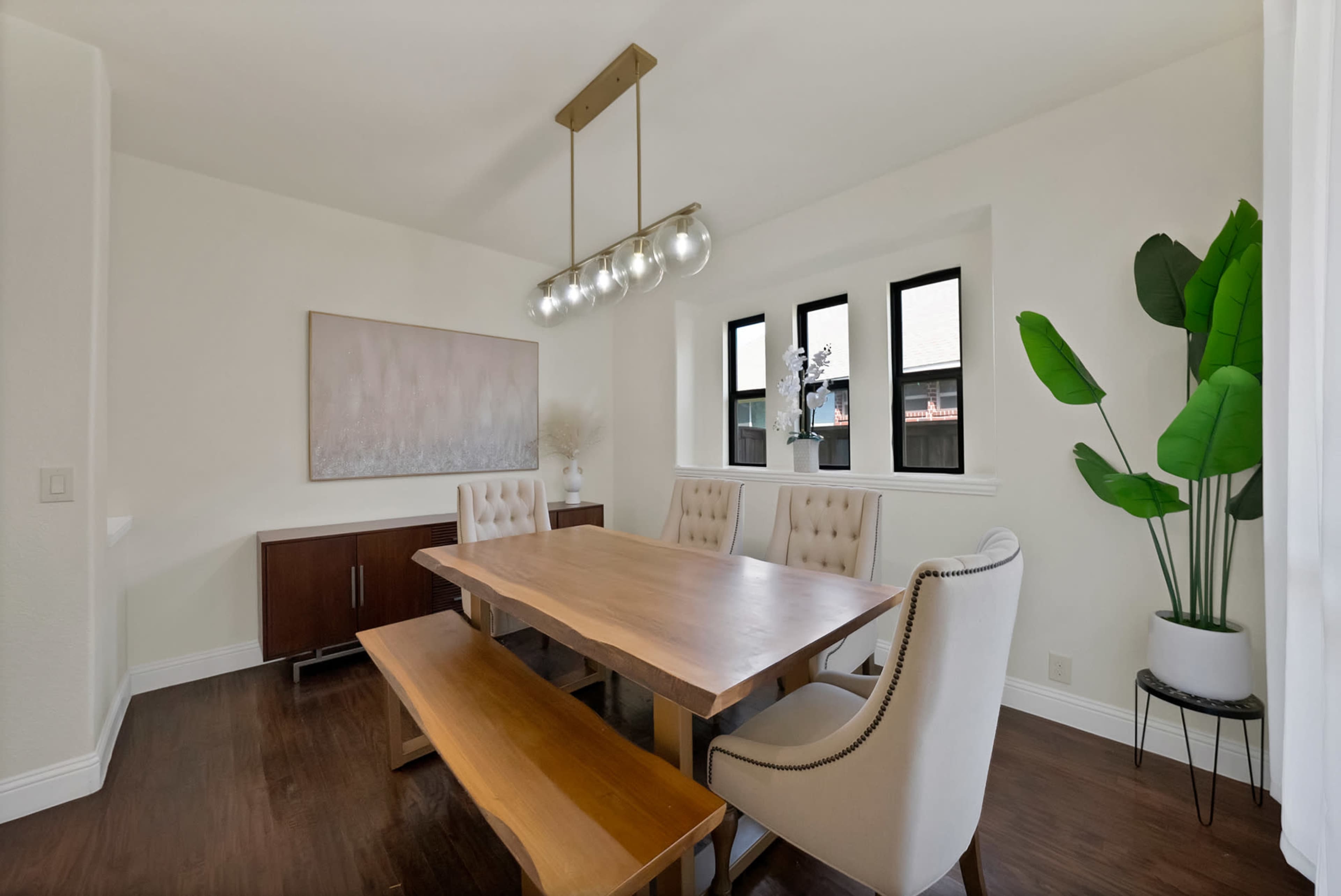 The image shows a modern dining room featuring a wooden table surrounded by beige upholstered chairs and a bench, with a large light fixture and a decorative plant in the corner.