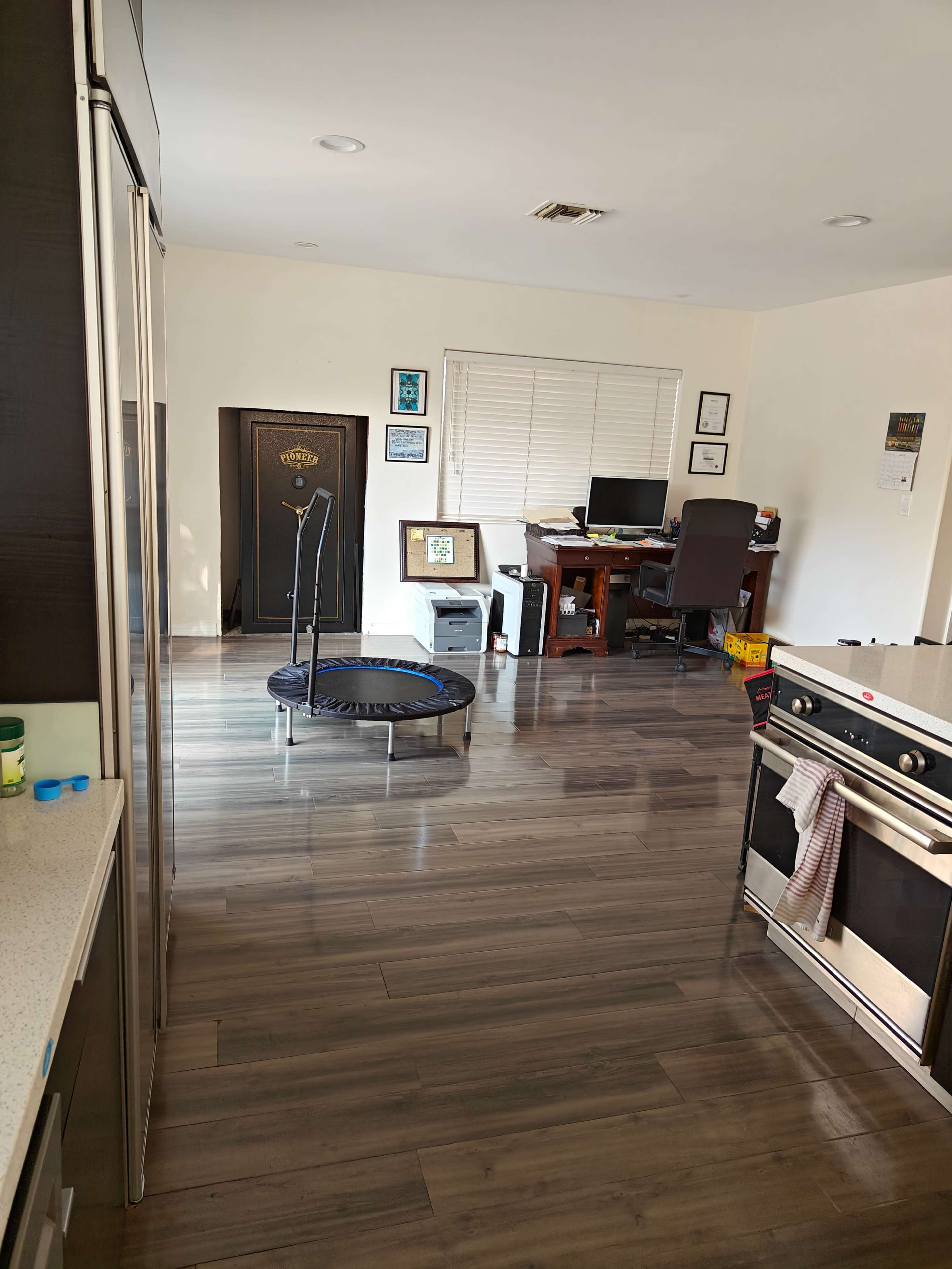 The image shows a modern kitchen area leading into a home office, featuring a small trampoline and a desk with a computer setup.