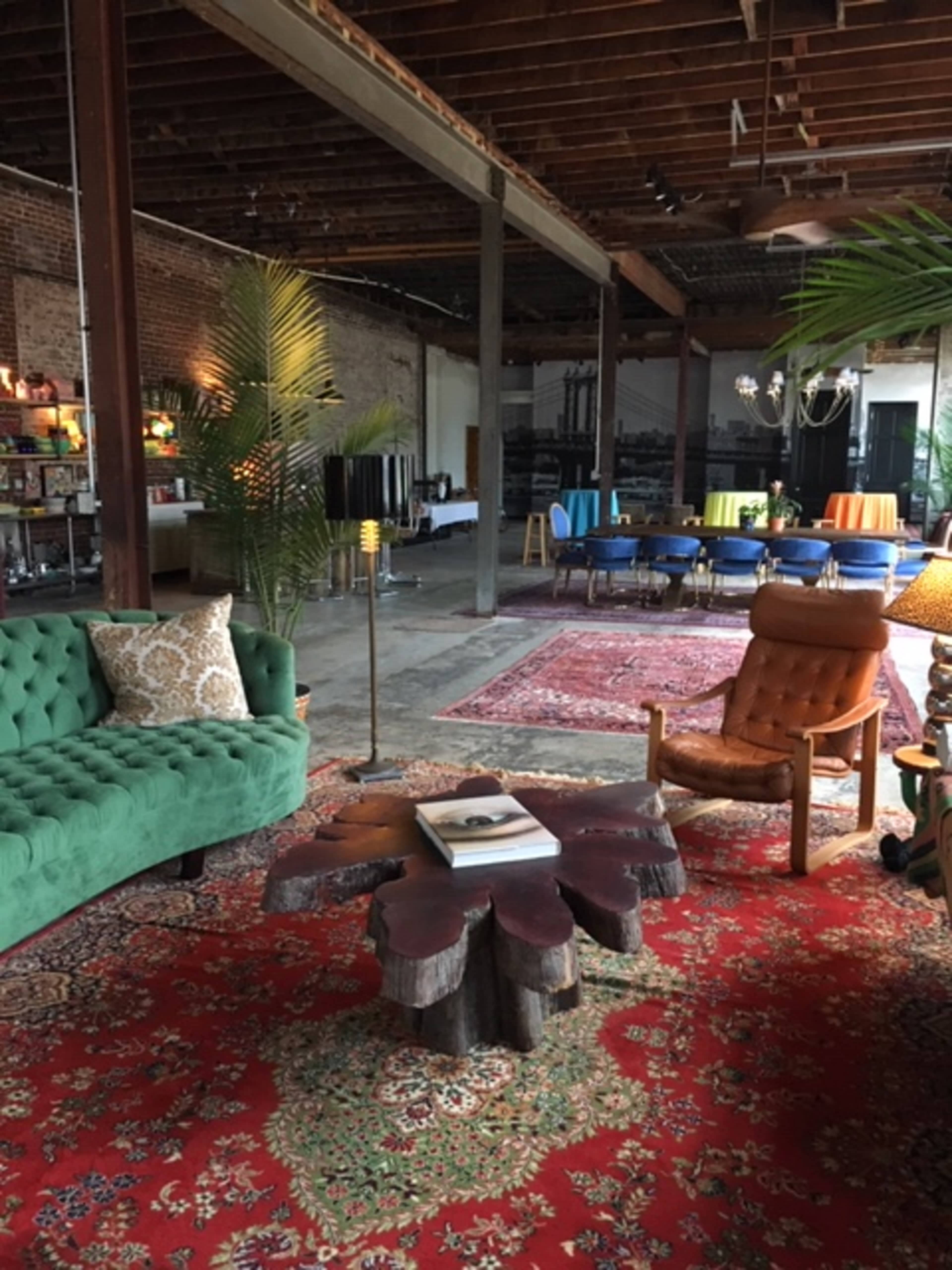 The image shows a spacious, industrial-style room featuring eclectic furniture including a green sofa, an orange armchair, and various tables, all arranged on a vibrant red patterned rug.