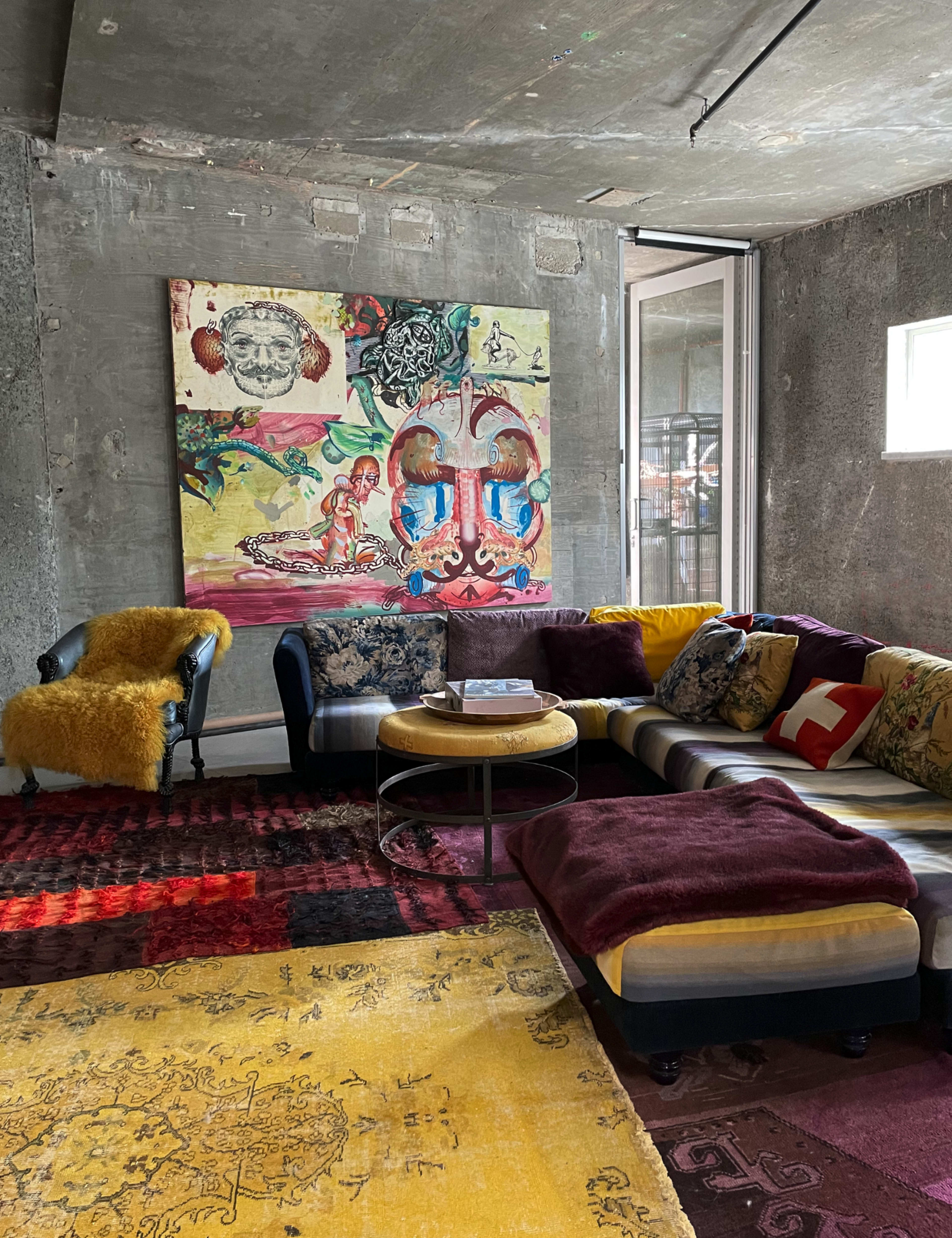The image shows a modern living room with a colorful sofa, various patterned cushions, an artistic wall mural, and a round coffee table on a mix of rugs.