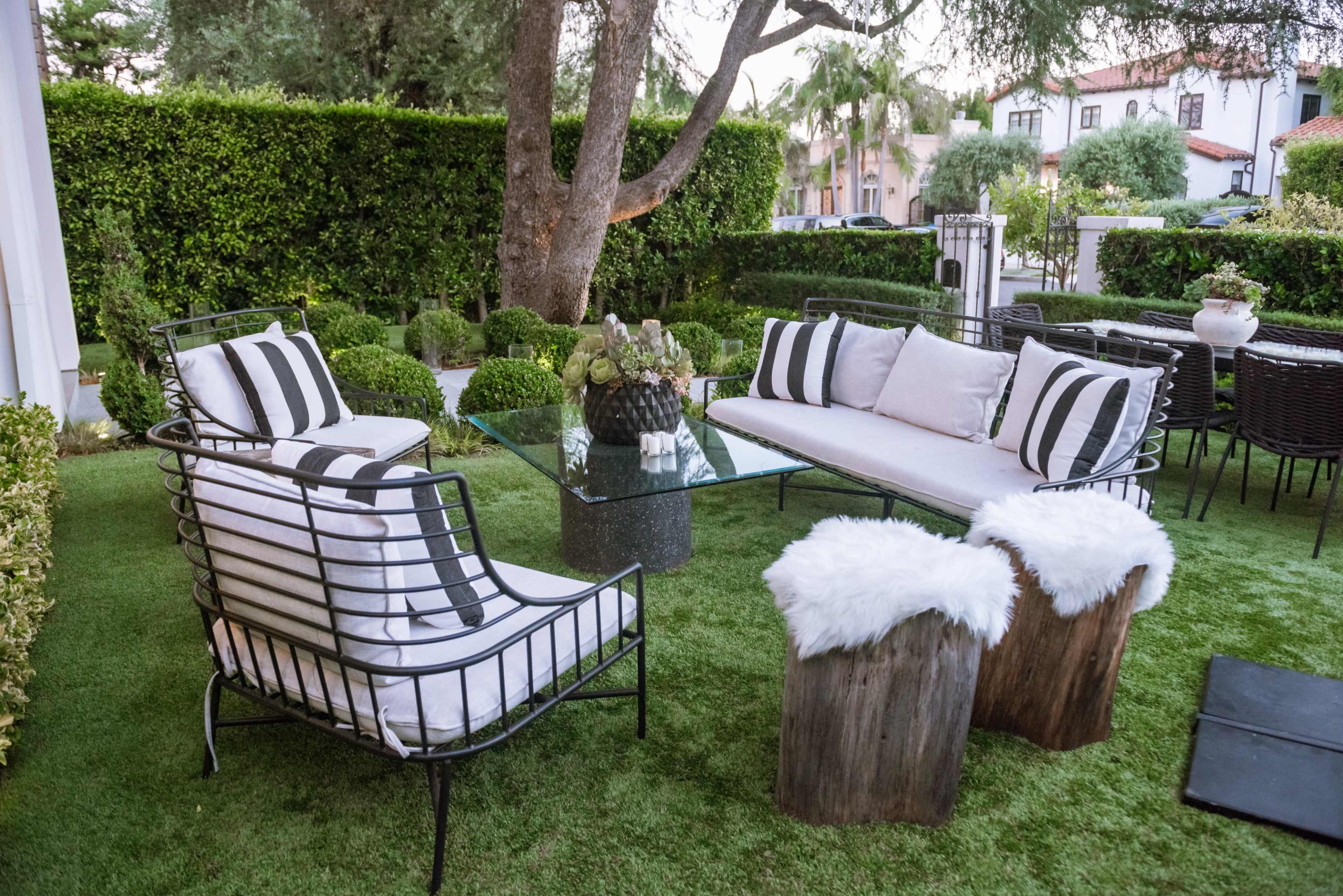 A modern outdoor seating area features black and white striped cushions on wire furniture, accented by a glass table and wooden stools, surrounded by manicured shrubbery and palm trees.