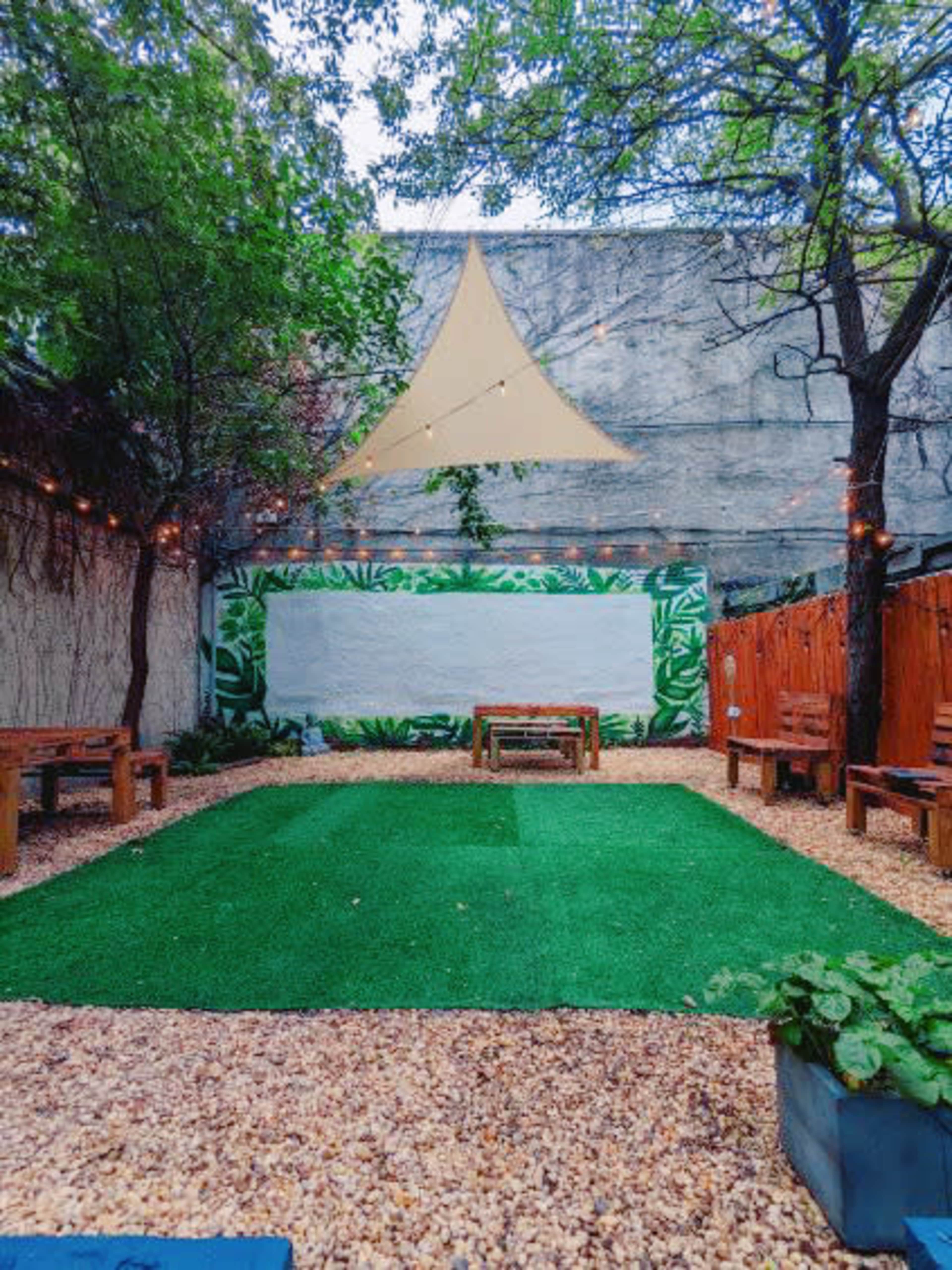 The image shows a small outdoor space with artificial grass, wooden benches, and a mural of greenery along one wall, all under a triangular canopy.