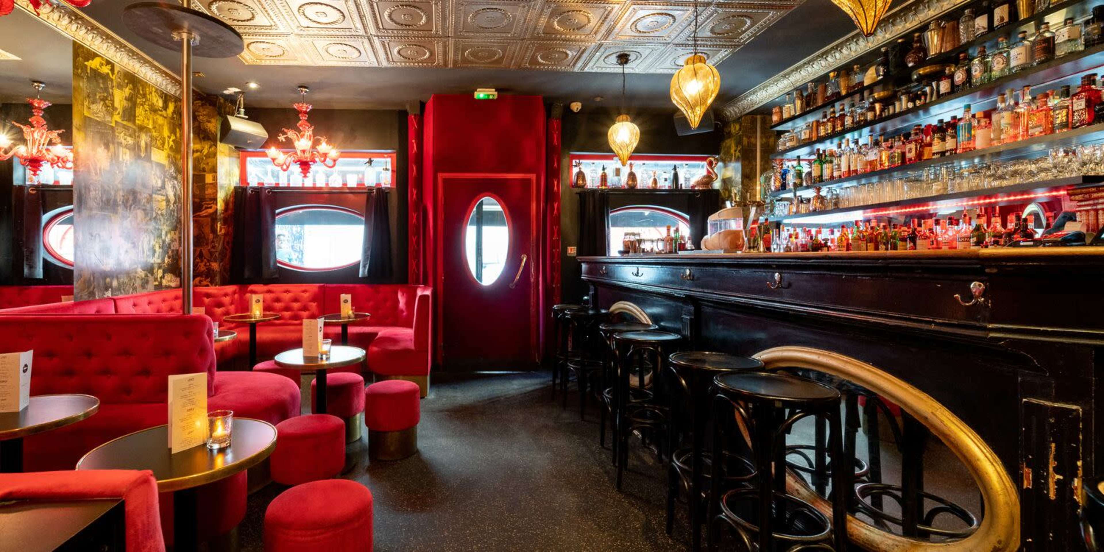 A stylish bar with red velvet seating, a dark wooden bar top, and shelves filled with various bottles of liquor under pendant lighting.