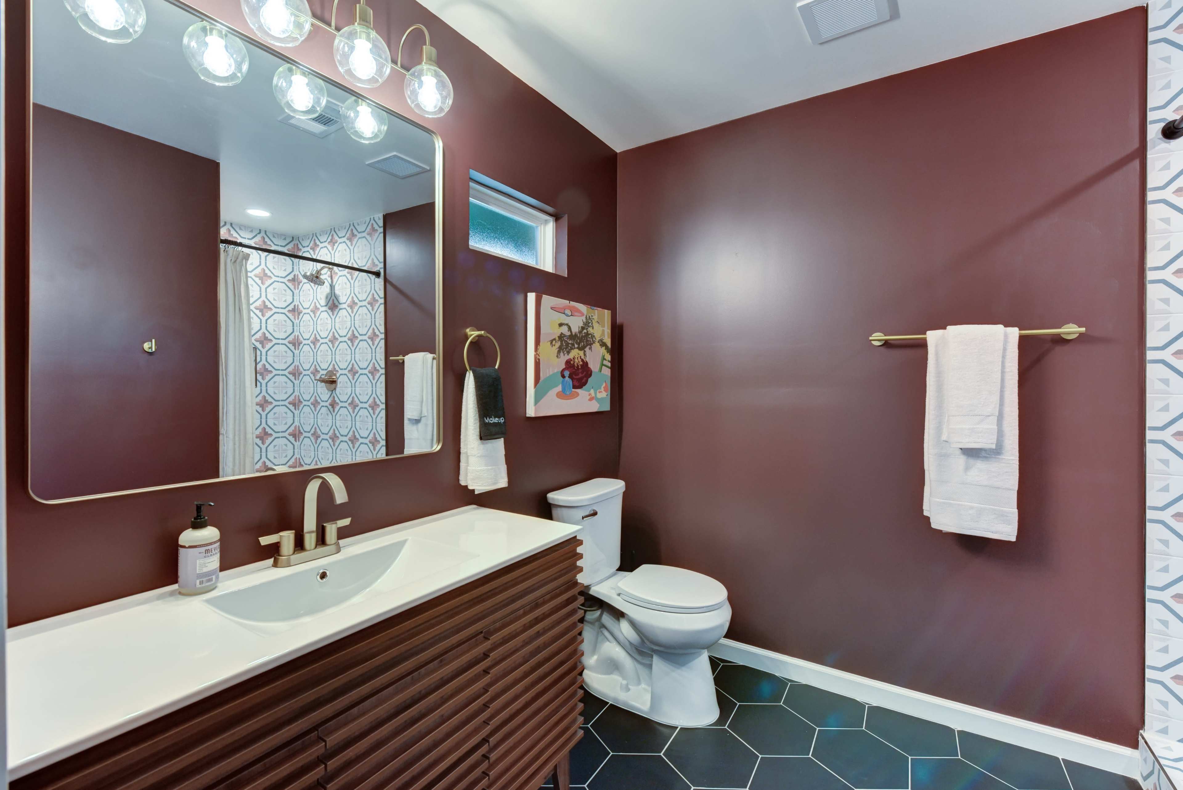The image shows a modern bathroom featuring a dark maroon wall, a large mirror with decorative lighting, a white vanity with brown cabinetry, a toilet, and patterned shower curtains.