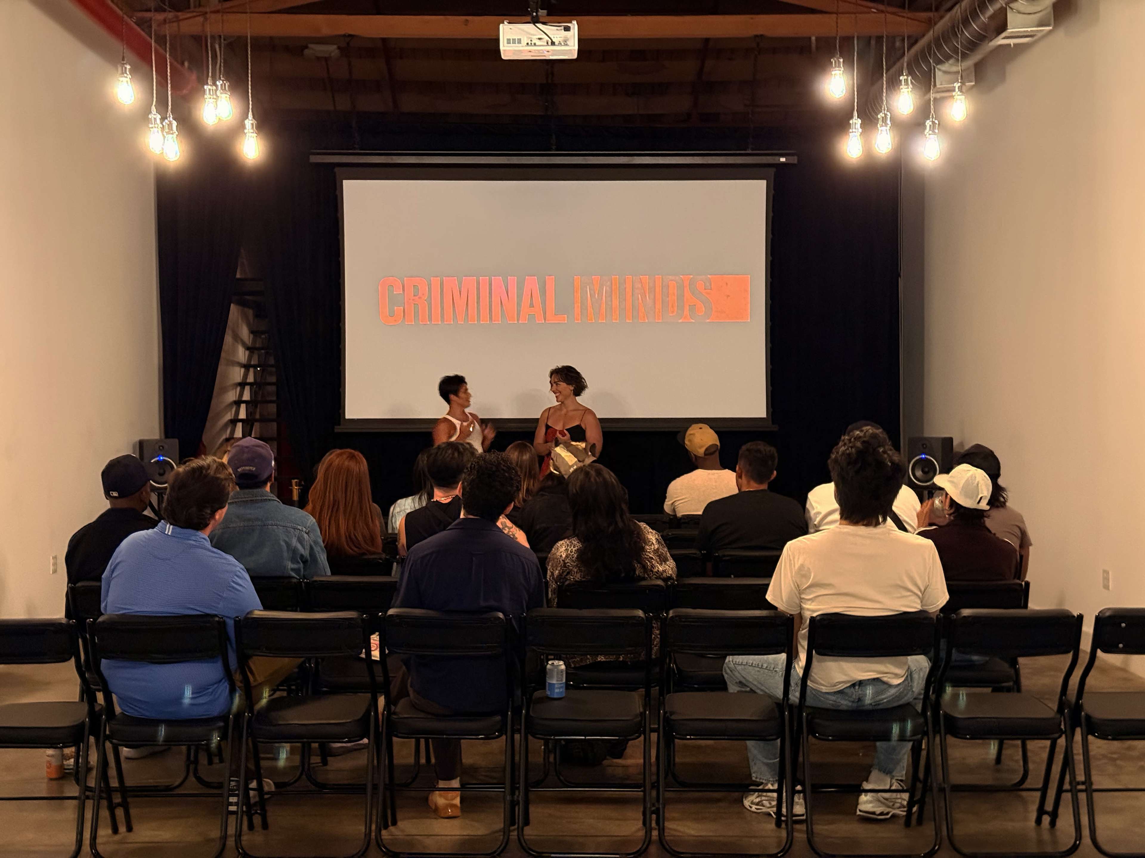 A small audience sits in folding chairs facing a large screen displaying the title "CRIMINAL MINDS," while two speakers engage in conversation on stage.