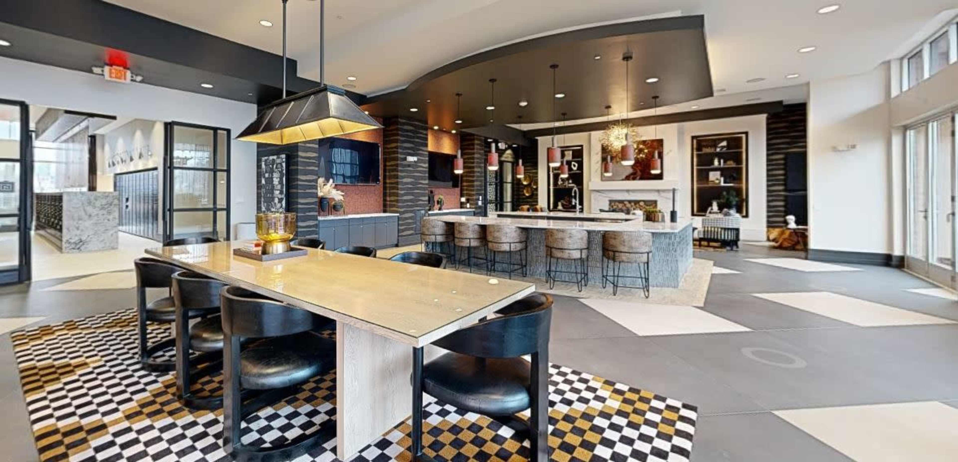 The image shows a modern lounge area featuring a large table with black chairs, a kitchen bar with stools, and a decorative fireplace in an open space.