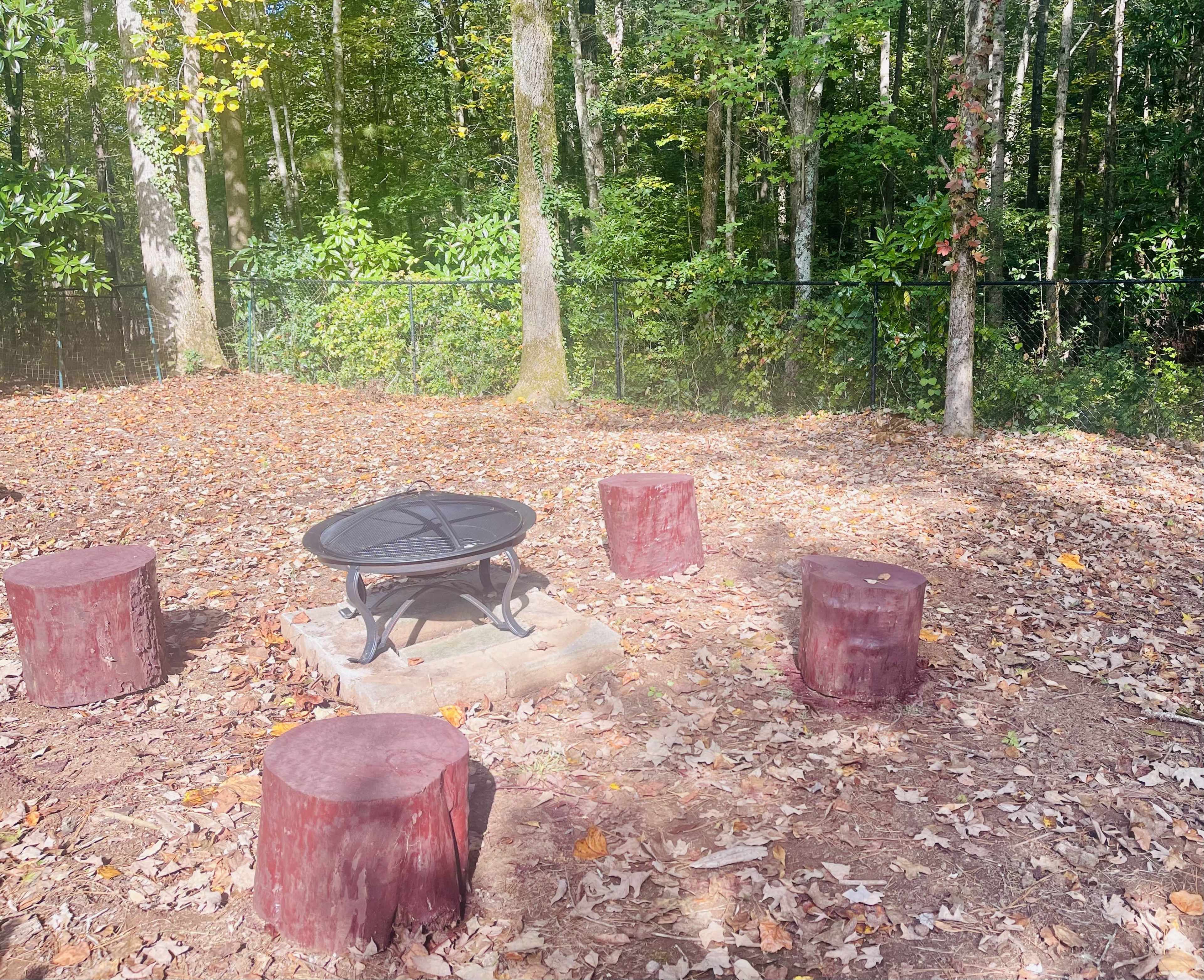 A circular fire pit surrounded by five log stools is situated in a wooded area covered with autumn leaves.