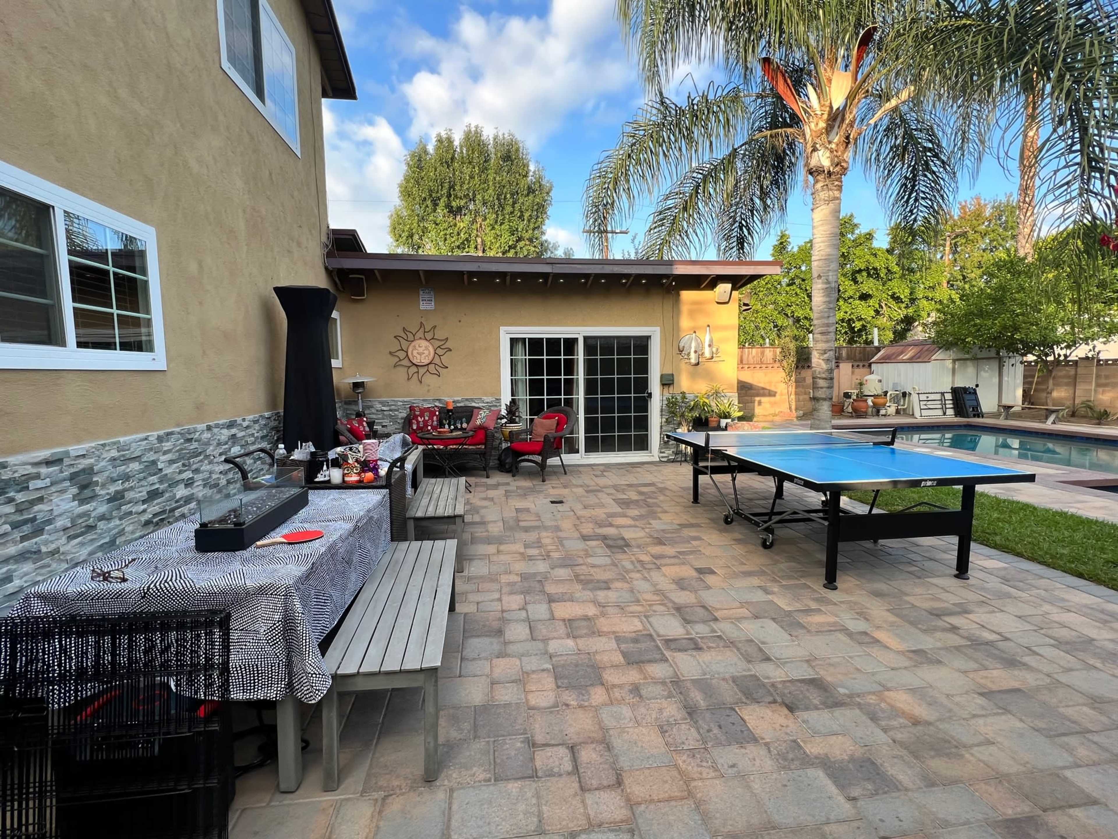 A backyard patio with a table set for dining, a ping pong table, and a swimming pool surrounded by palm trees.