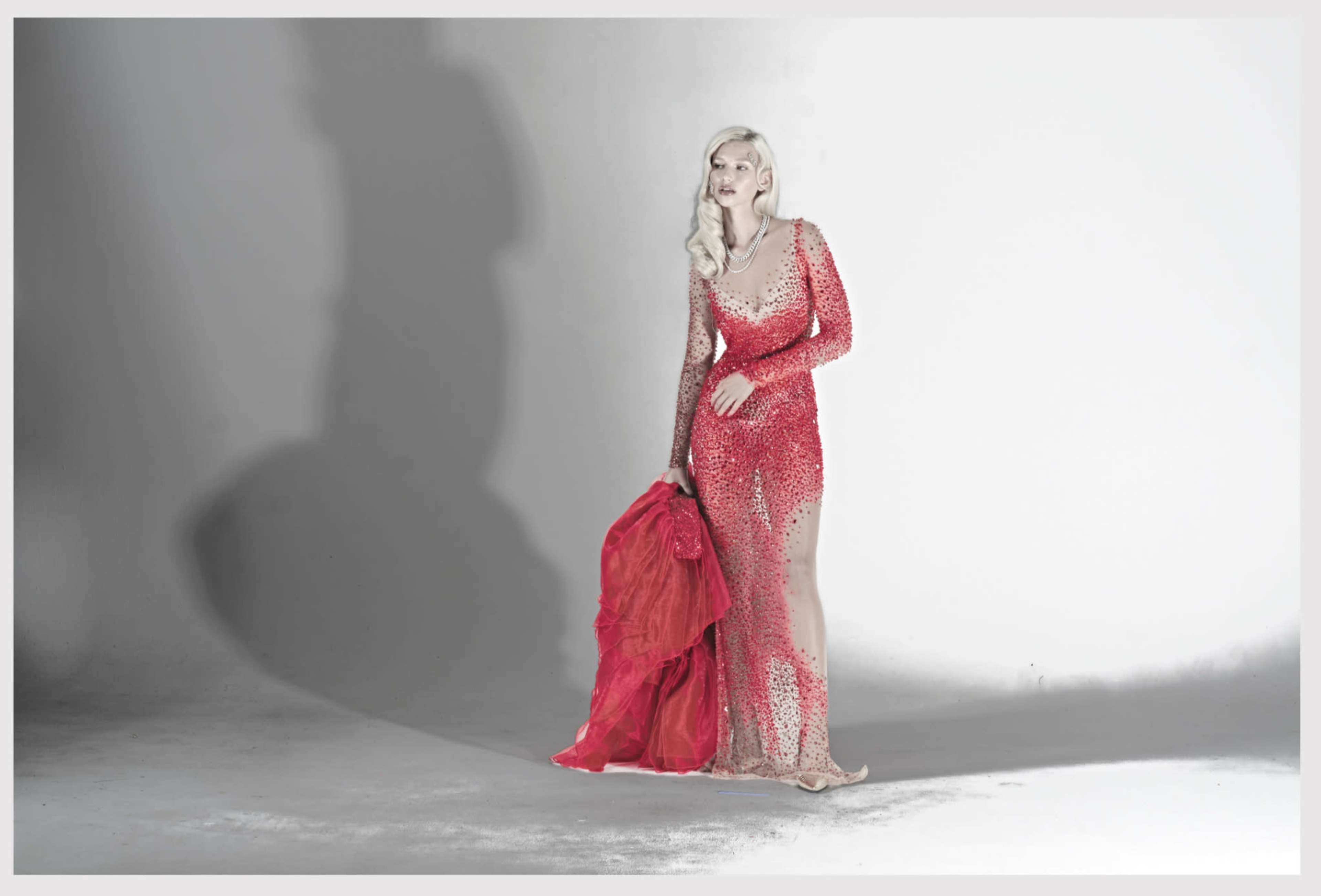 A model poses in a long, sparkly red gown, holding a matching fabric while standing against a plain white background.