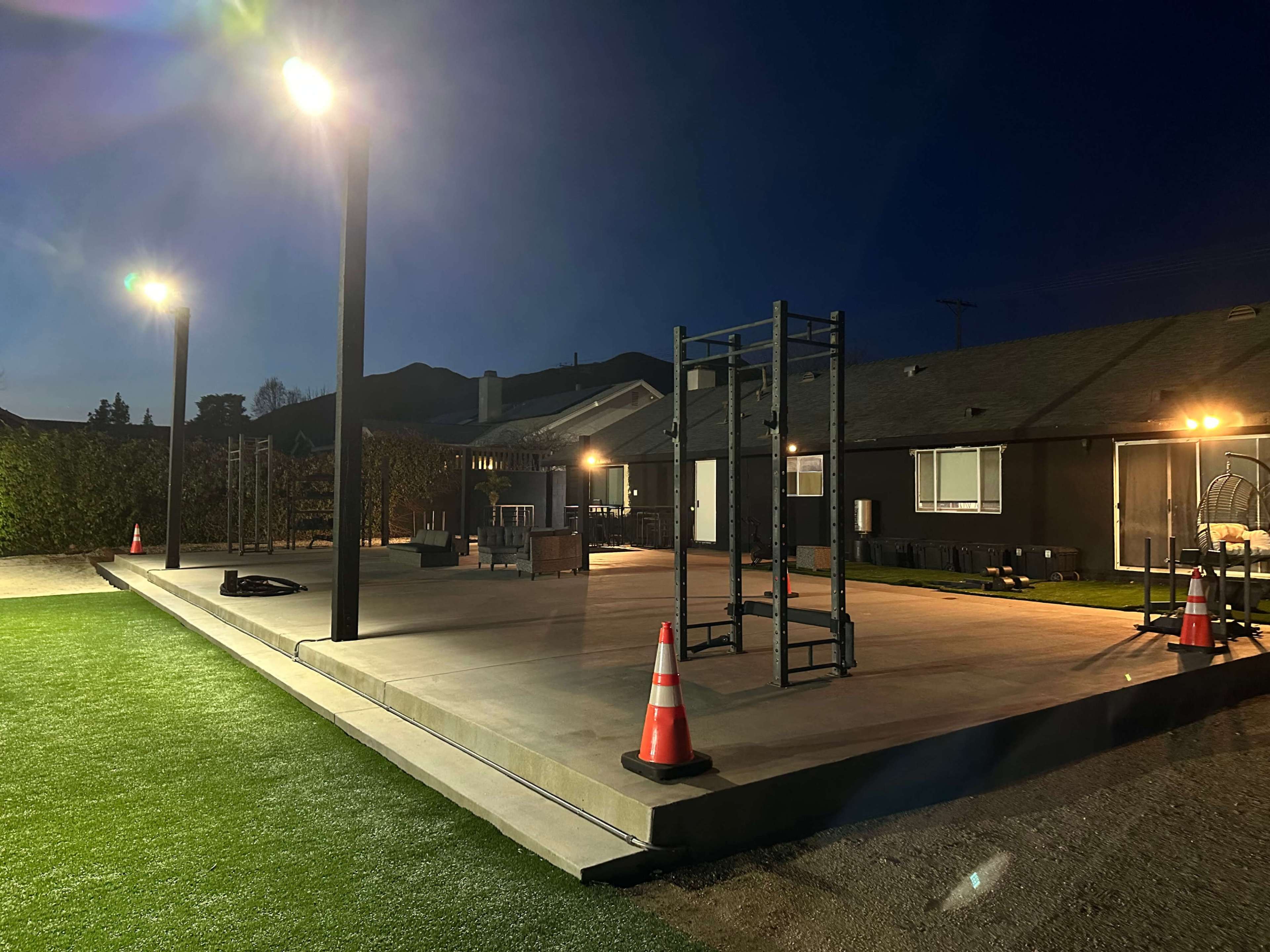 The image shows a flat, illuminated outdoor area at night, featuring workout equipment and traffic cones, with buildings visible in the background.