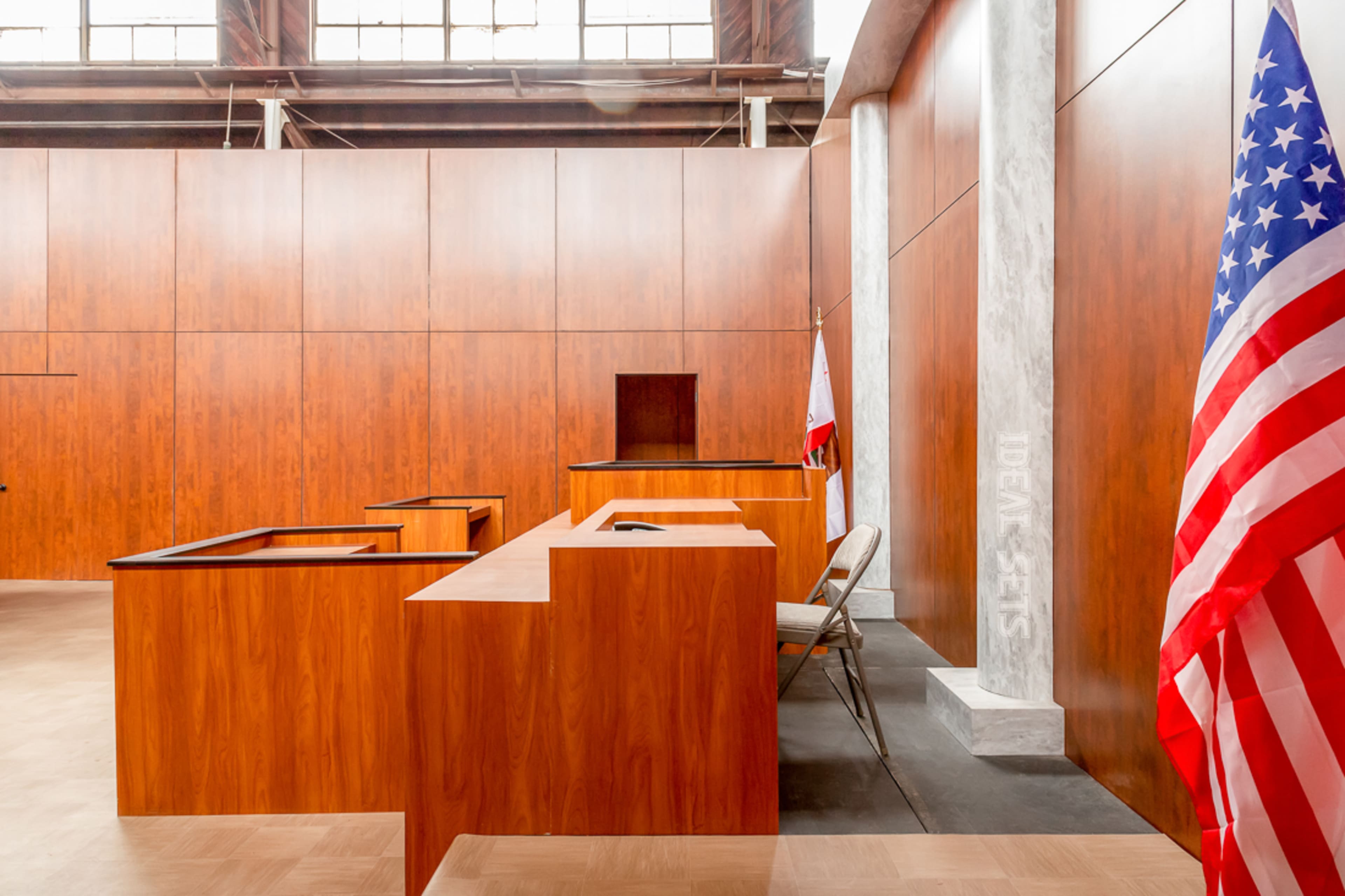 The interior of a courtroom with wooden paneling, empty witness and jury boxes, and an American flag in the foreground.