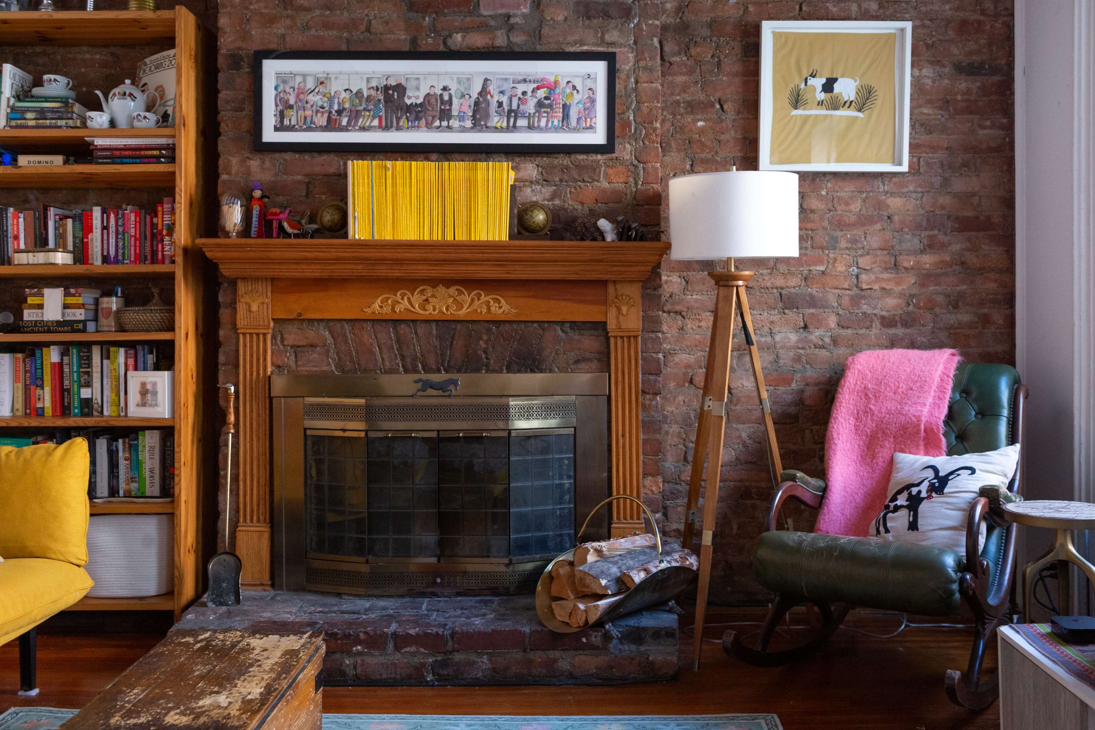 The image shows a cozy living room featuring a brick wall, a wooden fireplace mantel, a collection of books, a yellow armchair, and a green rocking chair with a pink throw blanket draped over it.