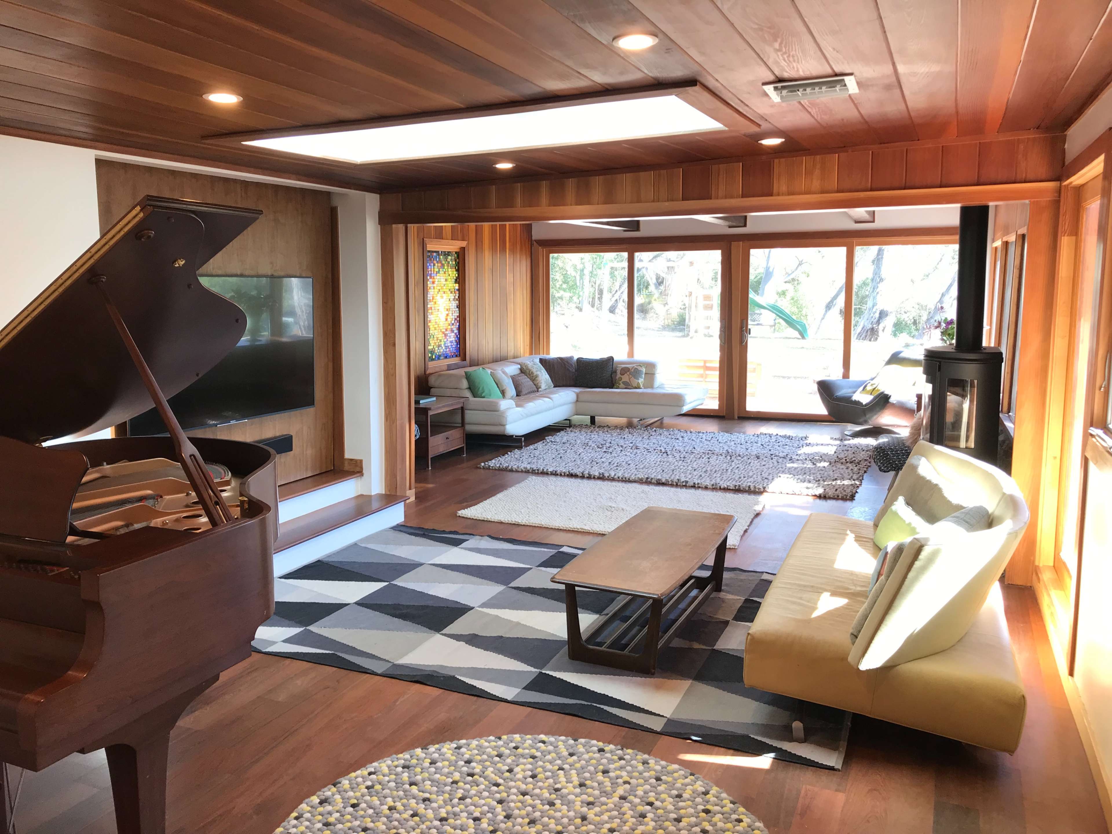 The scene shows a spacious living room with wooden paneling, a grand piano, a modern sofa arrangement, and large windows overlooking a landscaped outdoor area.