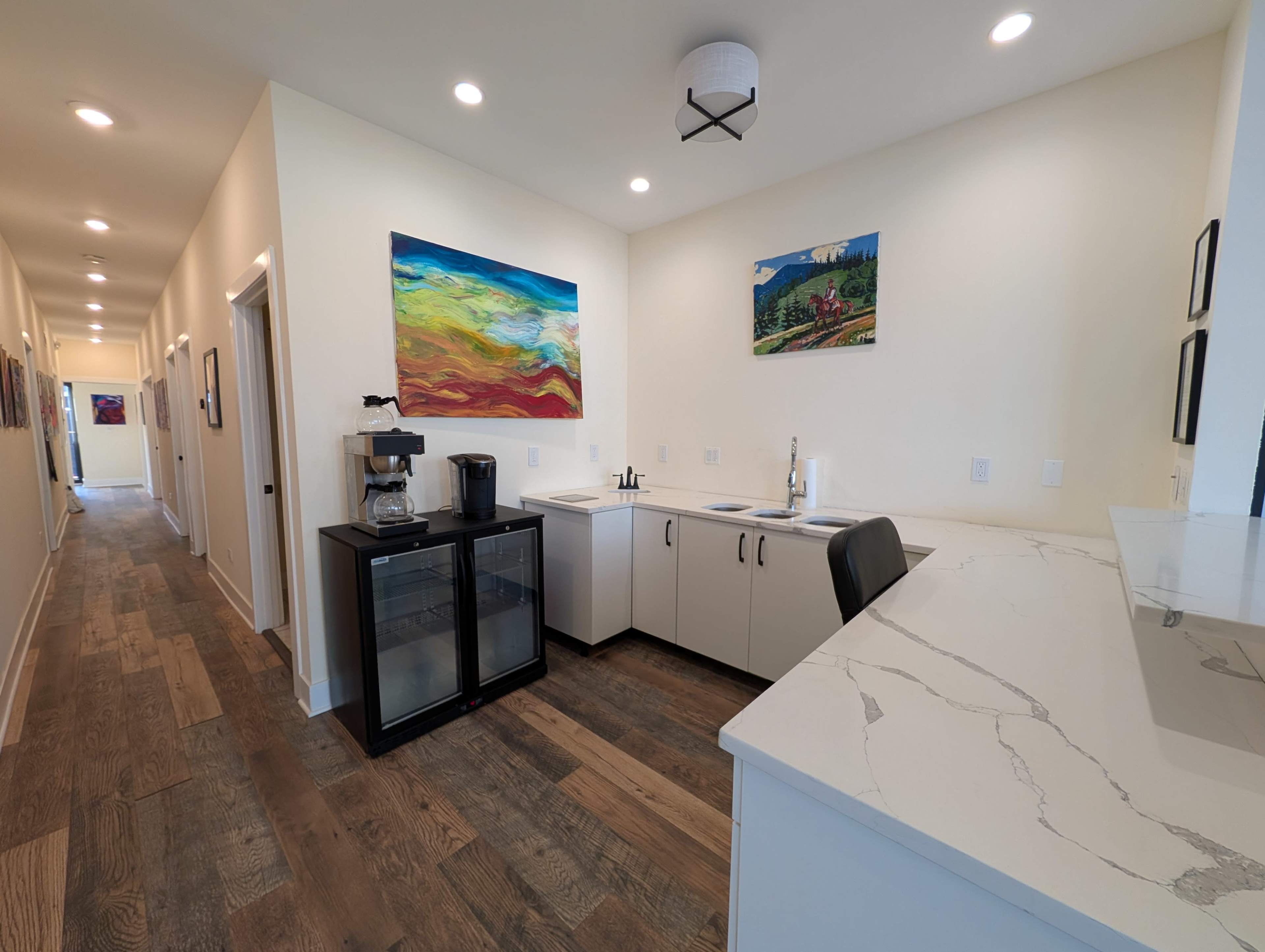 The image shows a hallway leading to a kitchenette with a coffee machine, a sink, and cabinets, alongside colorful artwork on the walls.