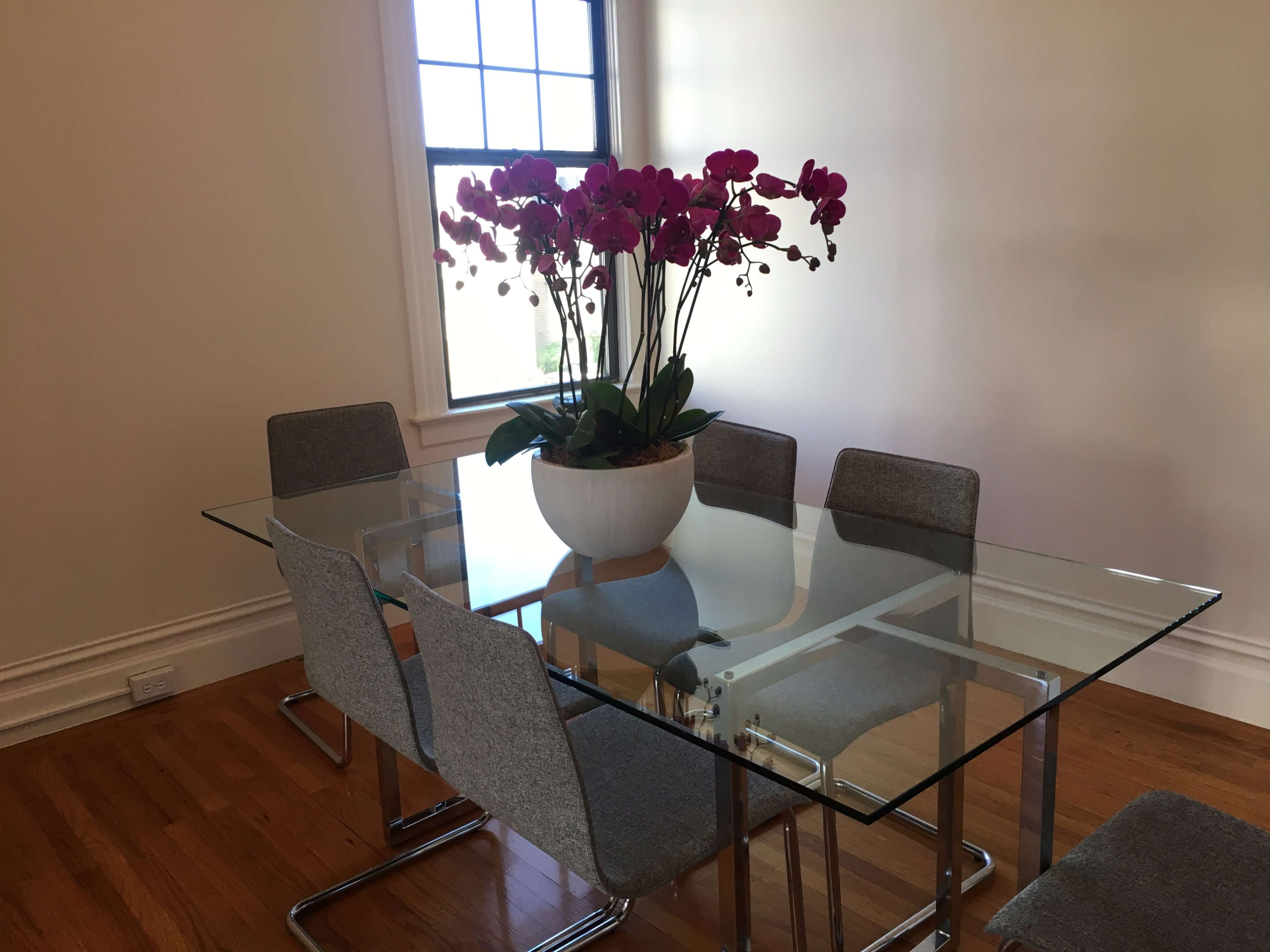 A glass dining table is set with six gray chairs and features a large white planter filled with pink orchids at its center.