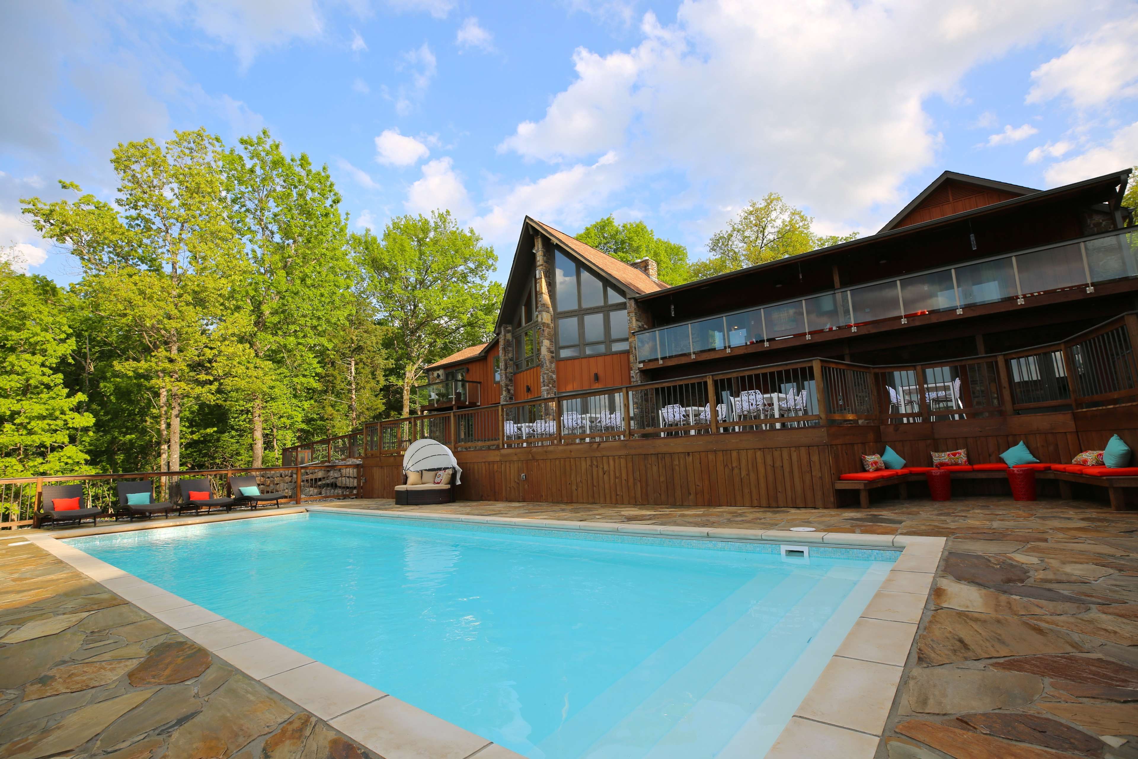 A swimming pool is located in front of a large wooden house surrounded by trees.