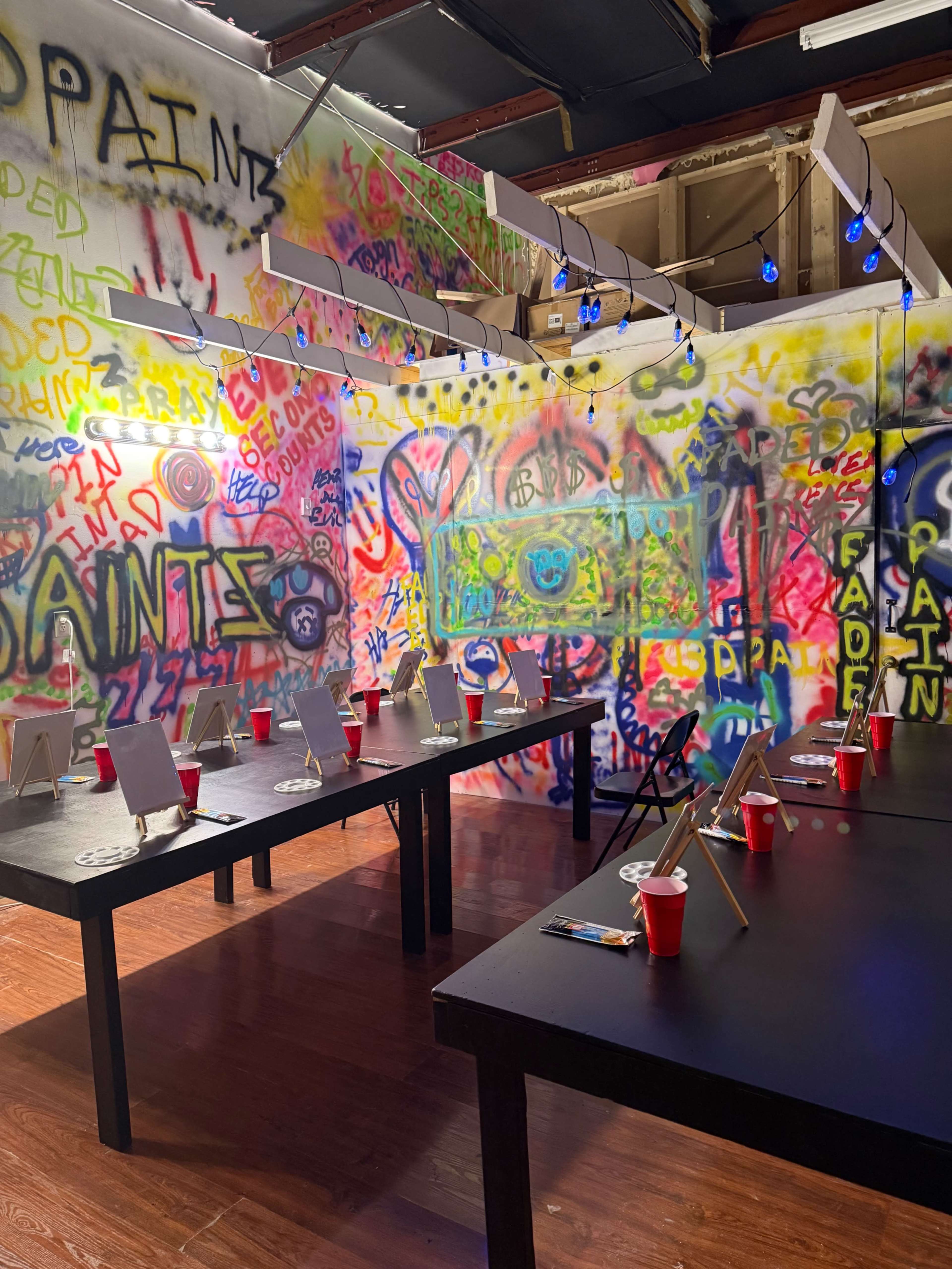 The image shows an artist studio with colorful graffiti-covered walls, several black tables, and red cups scattered around, along with small easels set up for painting.