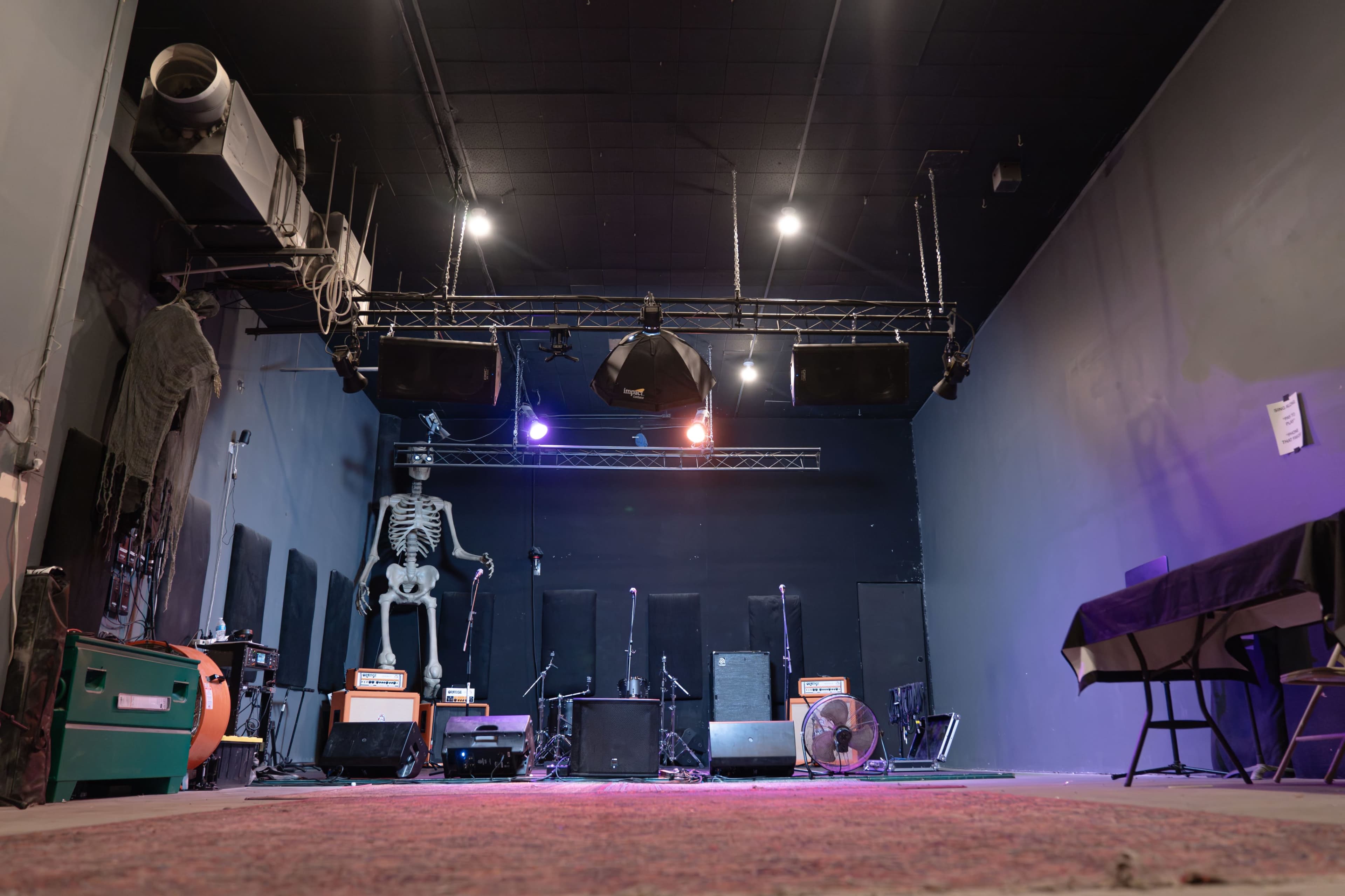 The image shows a dimly lit stage in a performance venue, featuring musical equipment, a skeleton prop, and an area with a carpet in front.