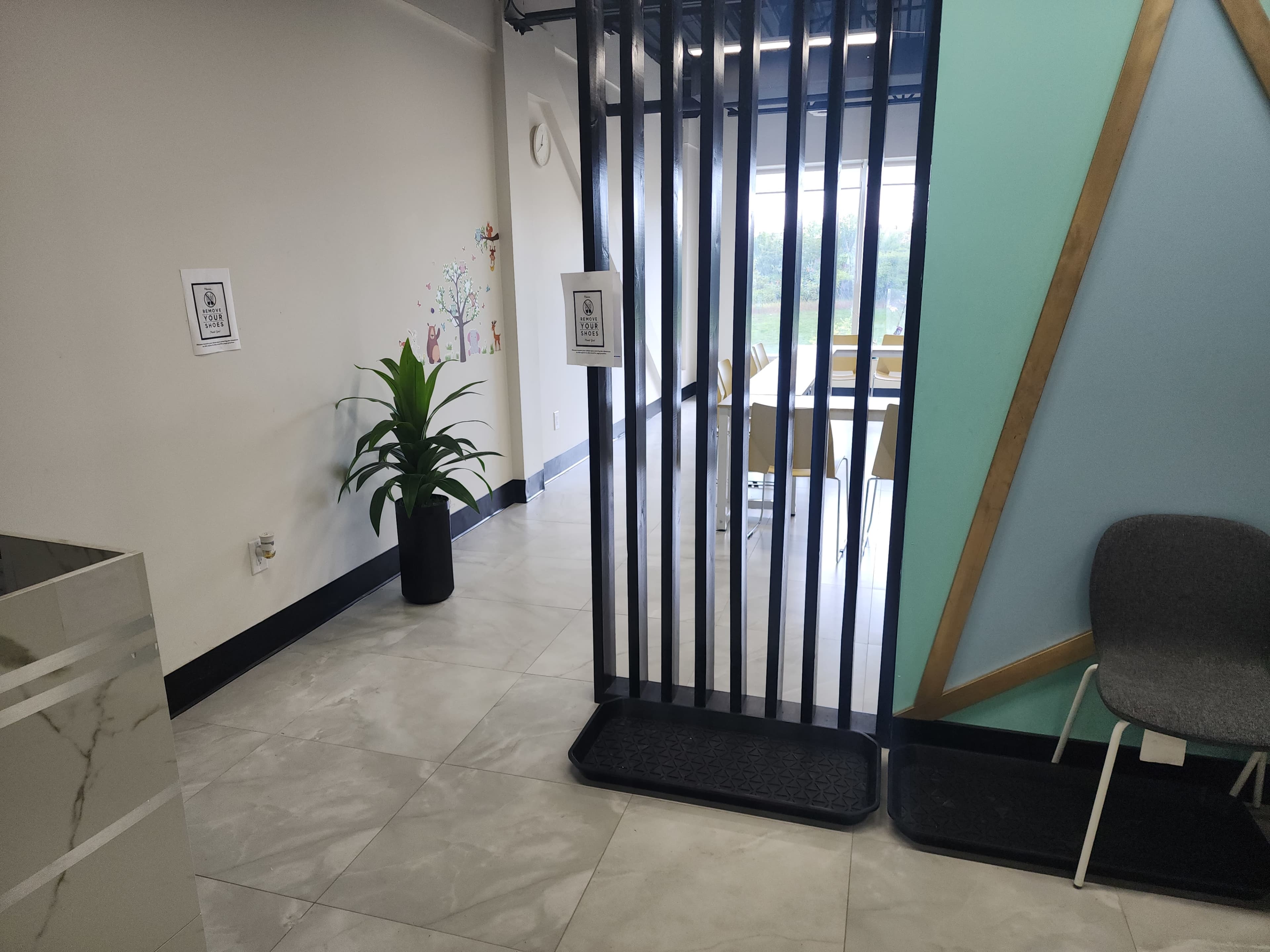 The image shows a modern indoor space with a partitioned area leading to a brighter room with tables and chairs, alongside a potted plant and a gray chair in the foreground.