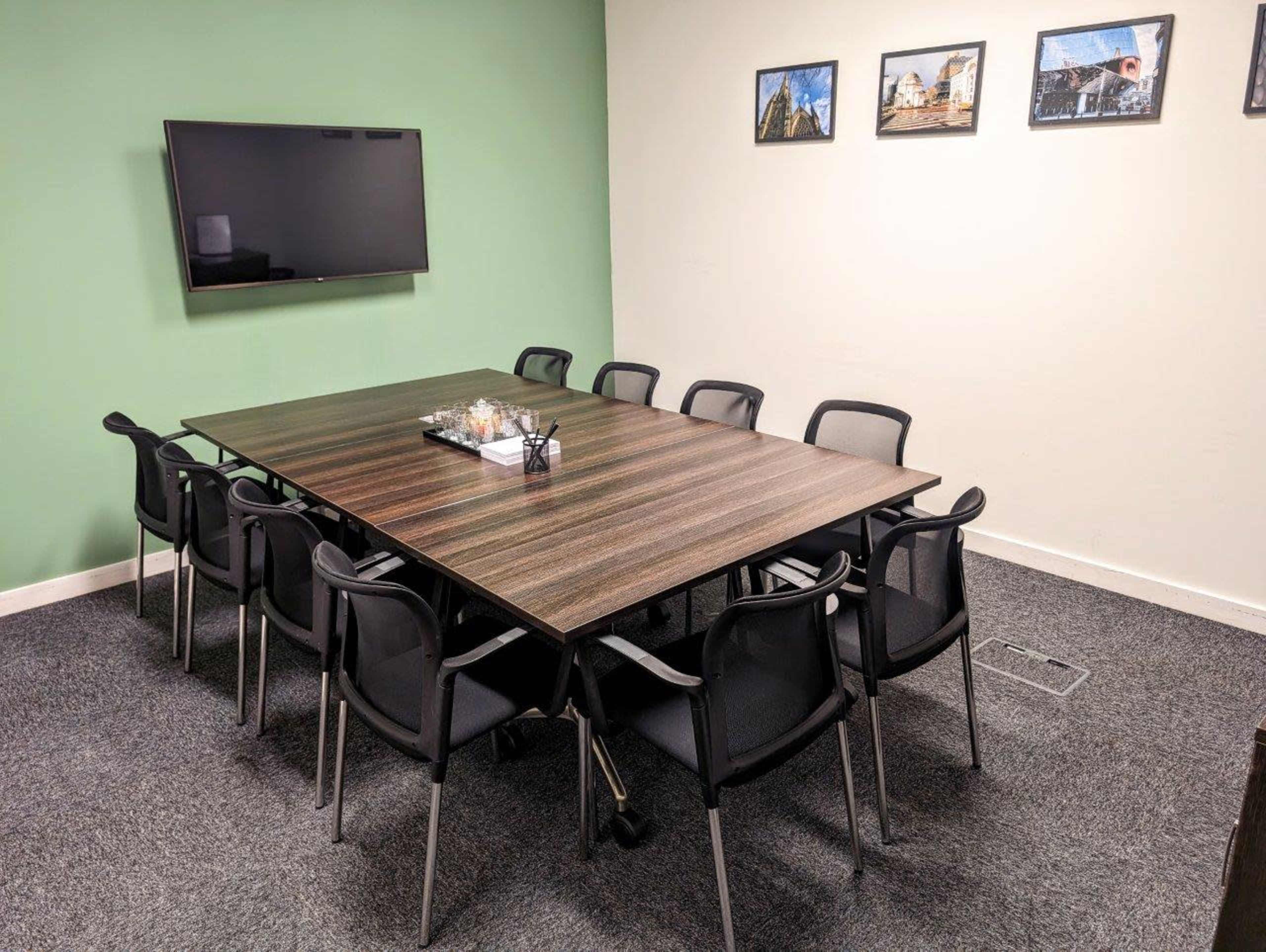 A conference room with a rectangular table surrounded by eight black chairs, a television mounted on the wall, and framed photographs hanging above.