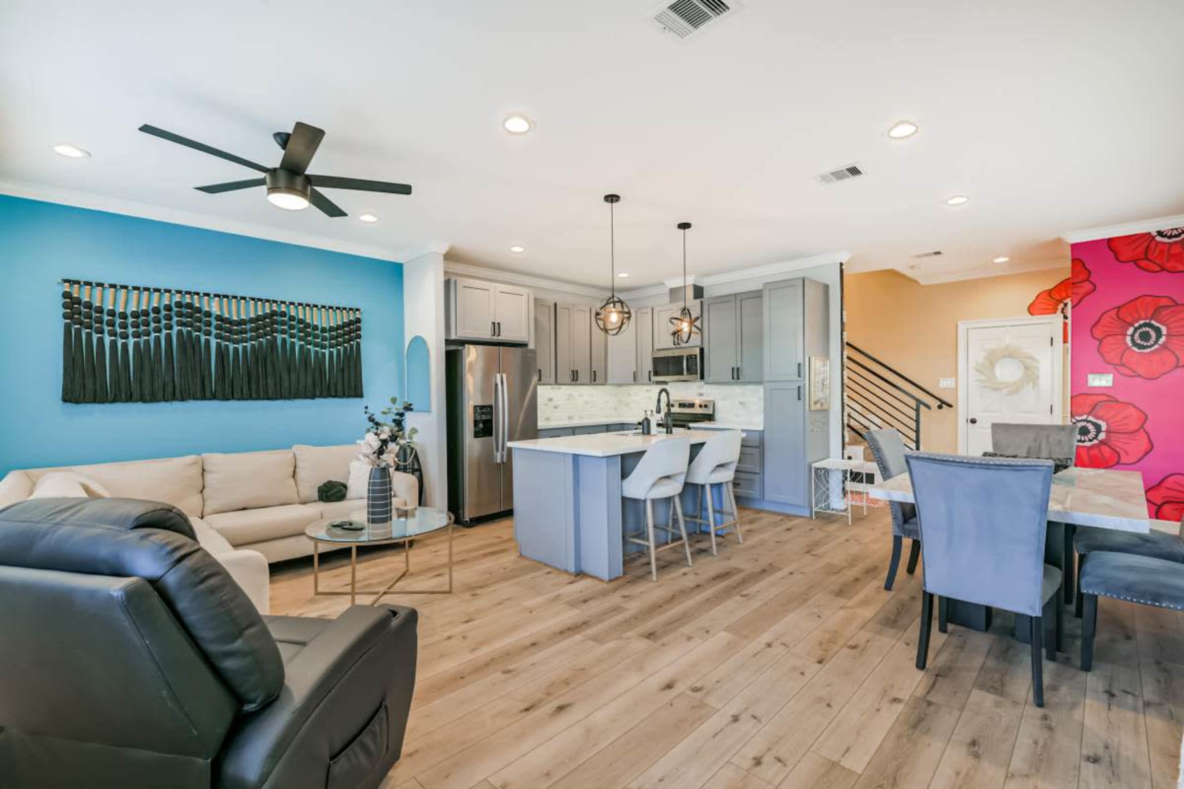 The image shows a modern living room and kitchen area featuring a light-colored sofa, a dining table, and blue and red accent walls with wooden flooring.