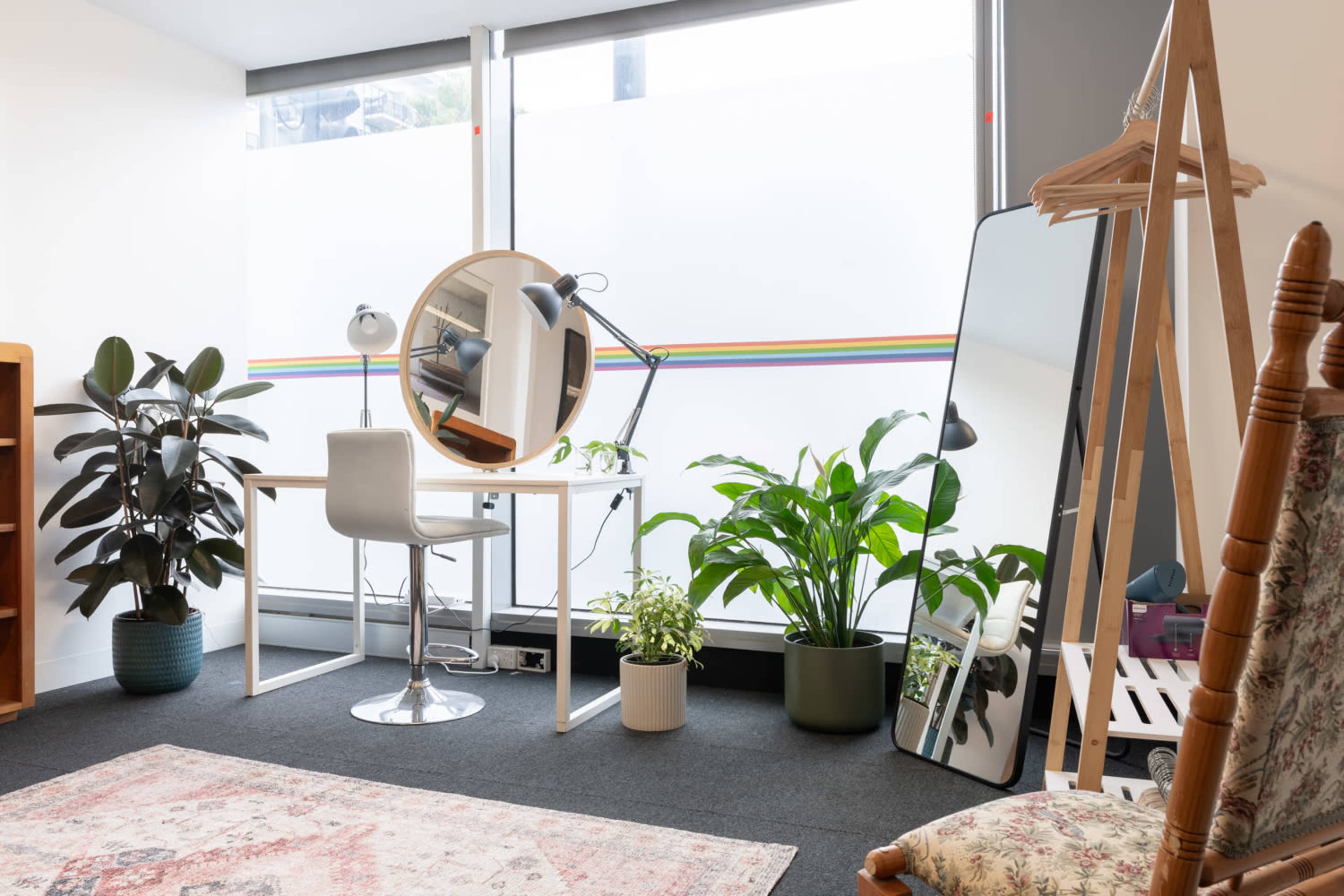 A well-lit room featuring a desk with a round mirror, a chair, various plants in pots, a large mirror, and a wooden clothing rack.